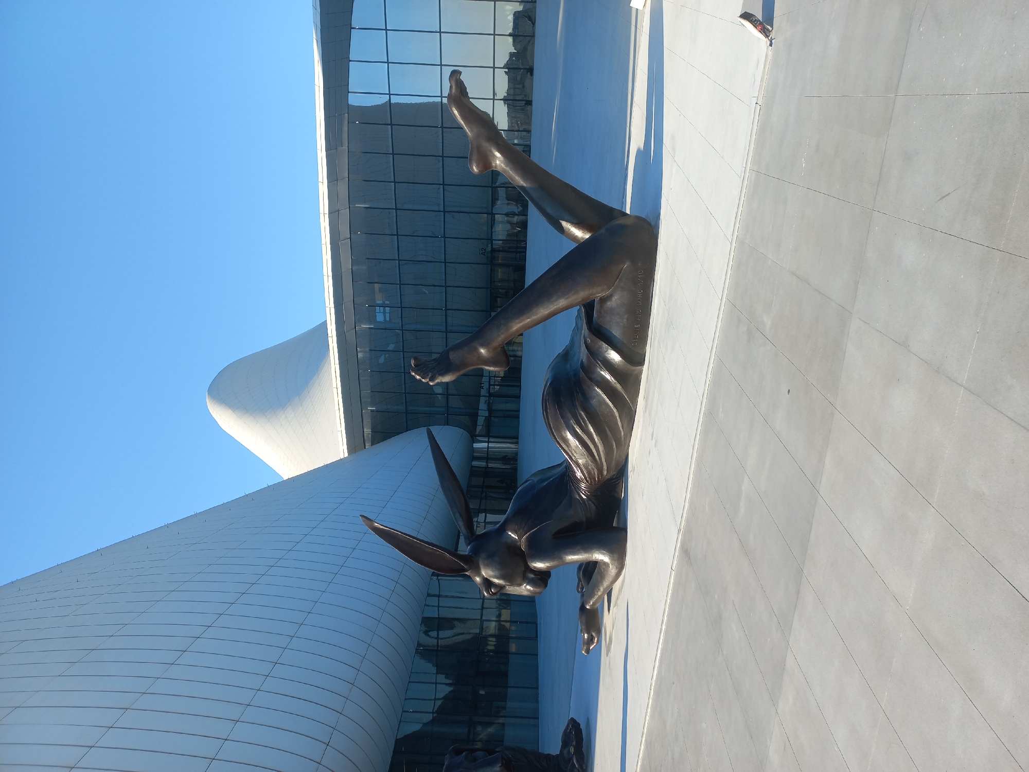 This image captures the exterior of the Heydar Aliyev Center in Baku, Azerbaijan, showcasing a large bronze sculpture of a reclining rabbit figure in the foreground. The sculpture, positioned on a paved plaza, depicts the rabbit in a relaxed pose, with its legs extended upwards. The modern, curvilinear architecture of the Heydar Aliyev Center forms a striking backdrop, characterized by its white, undulating facade and large glass panels. The sky above is clear and blue. To the far left of the image, a portion of another dark sculpture is visible. The scene is bathed in natural sunlight, casting shadows on the plaza. The inscription "ELLIE AND MARC 1/10" is visible on the base of the rabbit sculpture.