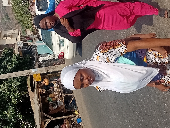 Two women stand on a paved road, with a car visible behind them. The woman in the foreground wears a white headscarf and patterned dress, while the other wears a black headscarf and bright pink outfit. A small stall or shop lines the road, overflowing with assorted items. It is located nearby a lush green tree.