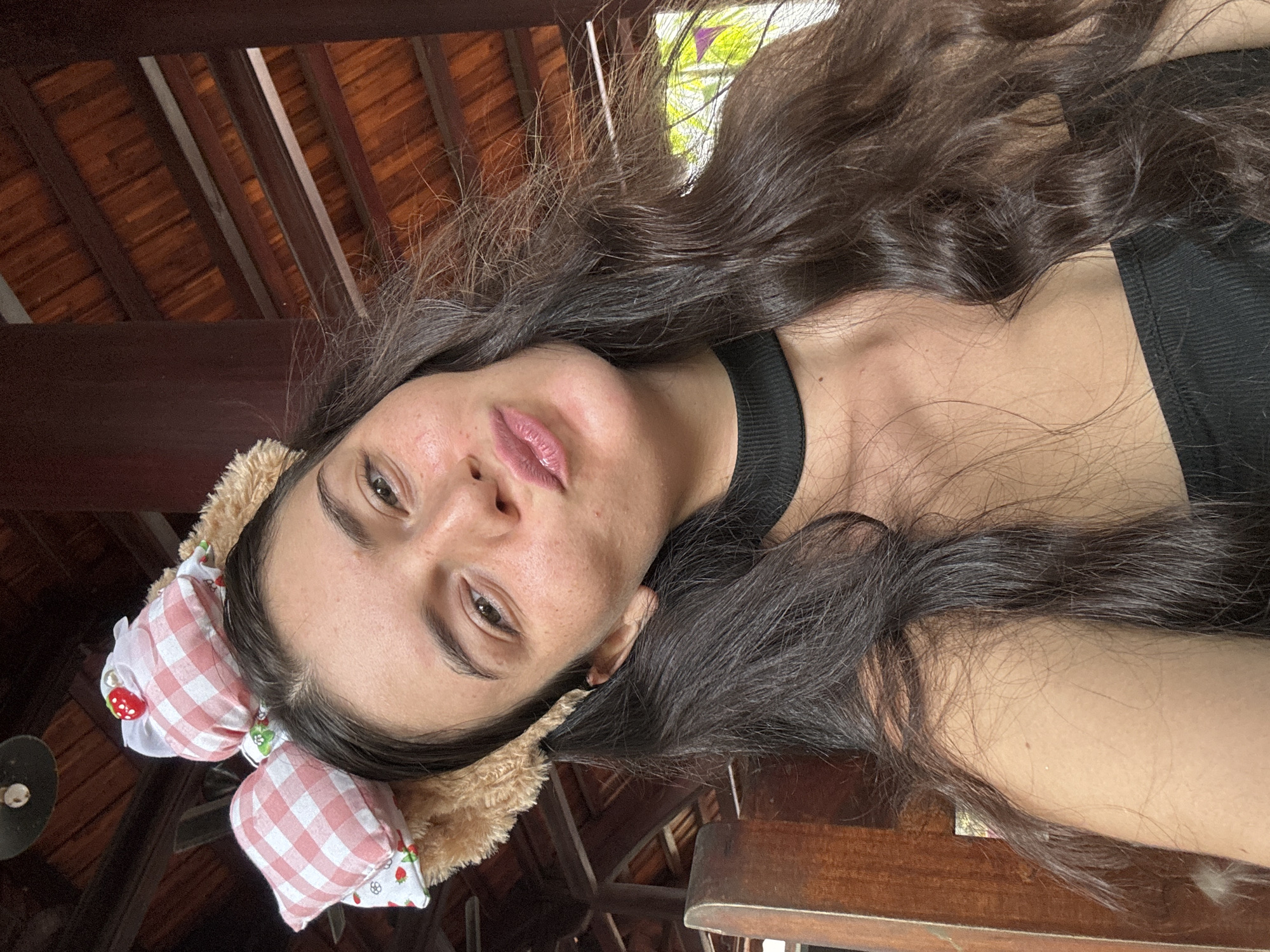 A young woman with long, dark, wavy hair is taking a selfie. She is wearing a black ribbed crop top and a black choker necklace. On her head, she has a headband with plush, tan, fuzzy ears and two pink and white checkered bows adorned with small red strawberry charms. Her lips are pursed, and she is looking directly at the camera. The background suggests an outdoor setting, possibly a cafe or restaurant with a wooden roof structure and beams visible. The overall ambiance is casual and playful. The location is identified as Mũi Né, Vietnam.