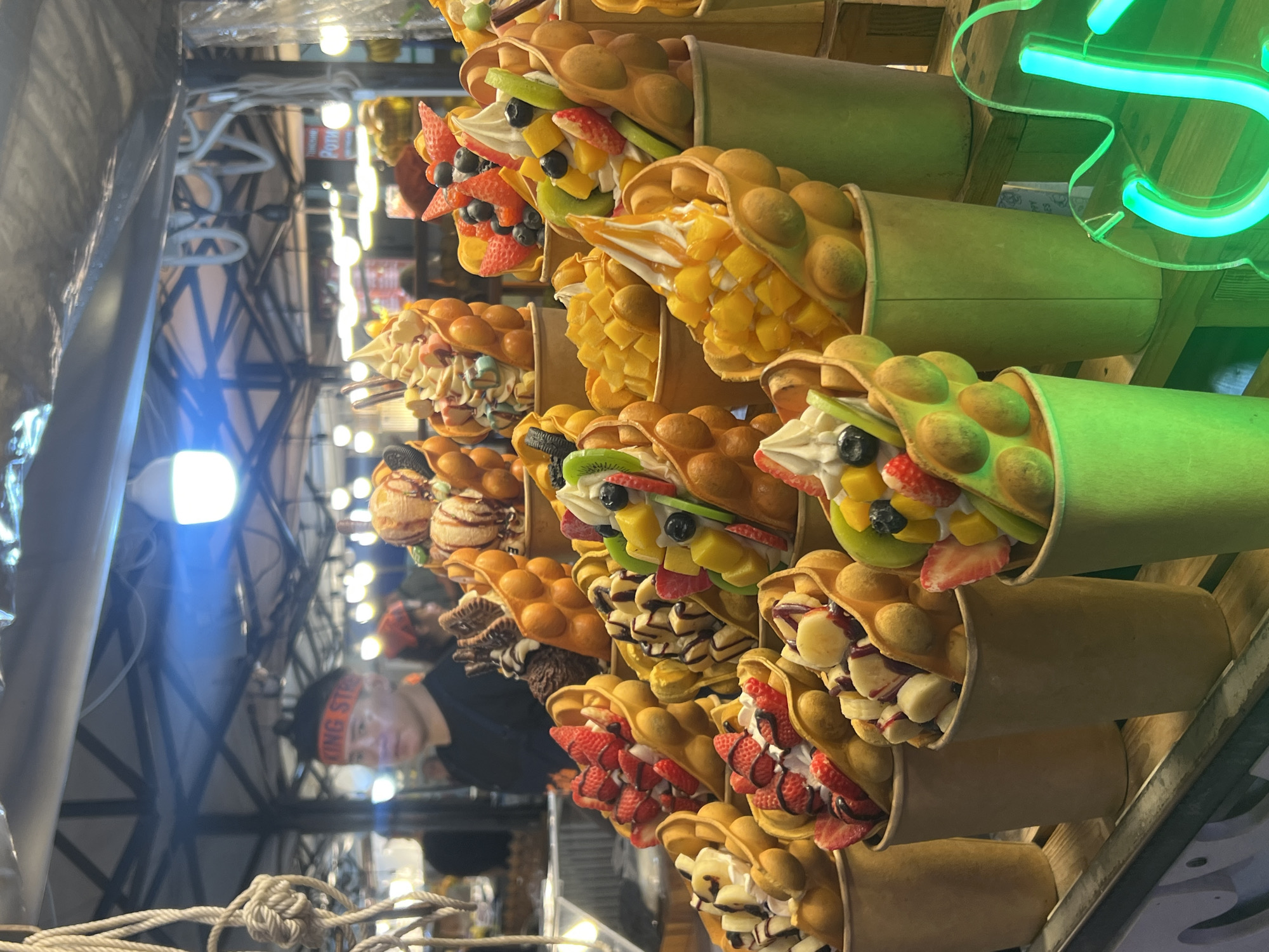The image shows a close-up of a street food stall in Karon, Thailand, selling bubble waffles. The stall is laden with various colorful bubble waffle creations, each presented in a paper cup. The waffles are filled with a combination of soft-serve ice cream, fresh fruits like strawberries, bananas, blueberries, mangoes, and kiwi, and drizzled with sauces. In the background, a vendor wearing a black shirt and an orange headband that reads "KING STREET" is visible. The stall appears to be set up under a tent with bright lights illuminating the scene, suggesting it is nighttime or a dimly lit indoor market. Neon signs are partially visible in the foreground, adding to the vibrant atmosphere. The overall impression is of a lively and tempting dessert stand.