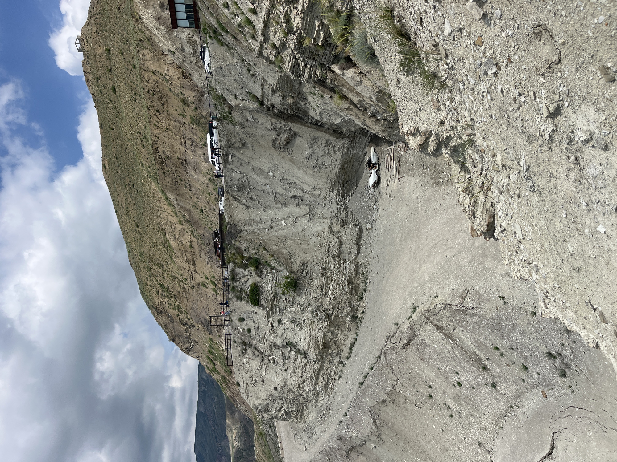 The image depicts a steep, rocky cliffside in Chirkey, Russia. A narrow road is carved into the cliff, with several cars parked along it. A metal walkway with railings runs parallel to the road, seemingly providing access to a building or viewing point at the top of the cliff. Two figures are visible near a vehicle at the edge of the cliffside, suggesting human activity. The environment is arid and mountainous, with limited vegetation. The sky is partially cloudy.