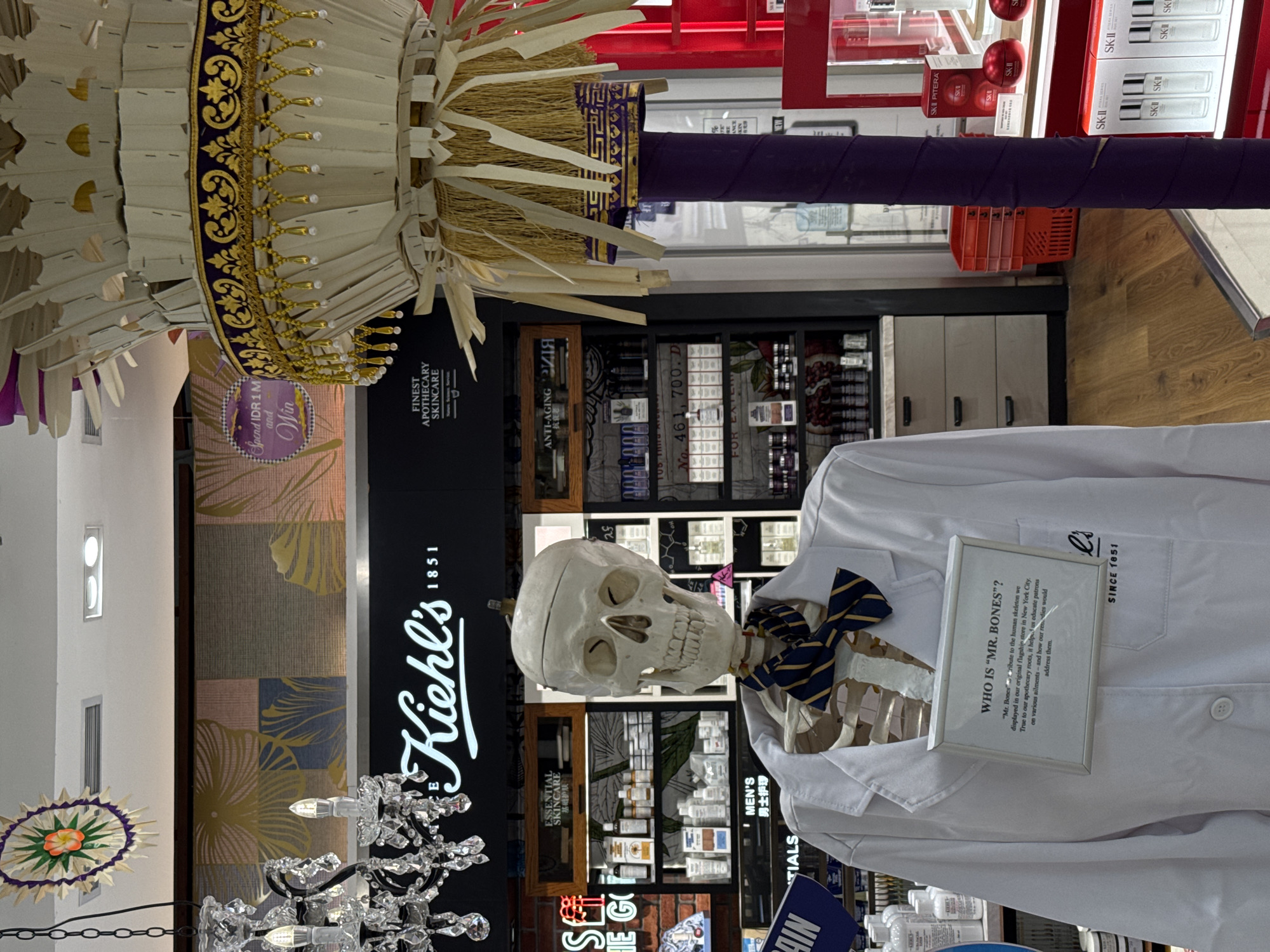 The image displays an indoor scene, likely a retail store, dominated by a life-sized skeleton figure named "Mr. Bones" standing on the right. Mr. Bones is wearing a white lab coat with "Kiehl's" and "SINCE 1851" visible on the pocket, and a blue and yellow striped bow tie. A framed sign hangs from his neck, explaining, "WHO IS 'MR. BONES'? 'Mr. Bones tribute to the human skeleton we displayed in our original flagship store in New York City. True to our apothecary roots, it helps us educate patrons on various ailments and how our remedies would address them."
Behind Mr. Bones and to the left, a "Kiehl's 1851" sign with "FINEST APOTHECARY SKINCARE" is prominently displayed on a dark background. Store shelves filled with various skincare products are visible, labeled with categories like "ANTI-AGING," "ESSENTIAL SKINCARE," and "MEN'S," some with Chinese translations. A promotional circular sign states "Spend IDR 1 M and Win."
In the upper left, a large, ornate decorative structure made of light-colored, possibly woven materials and straw-like elements, adorned with gold and purple accents, is partially visible. Below it, a hanging chandelier with clear crystal-like elements adds to the decor. Further to the right, a display of SK-II products in red and white packaging is seen on another shelf. The floor is light-colored wood. The overall setting is brightly lit, typical of a retail environment.