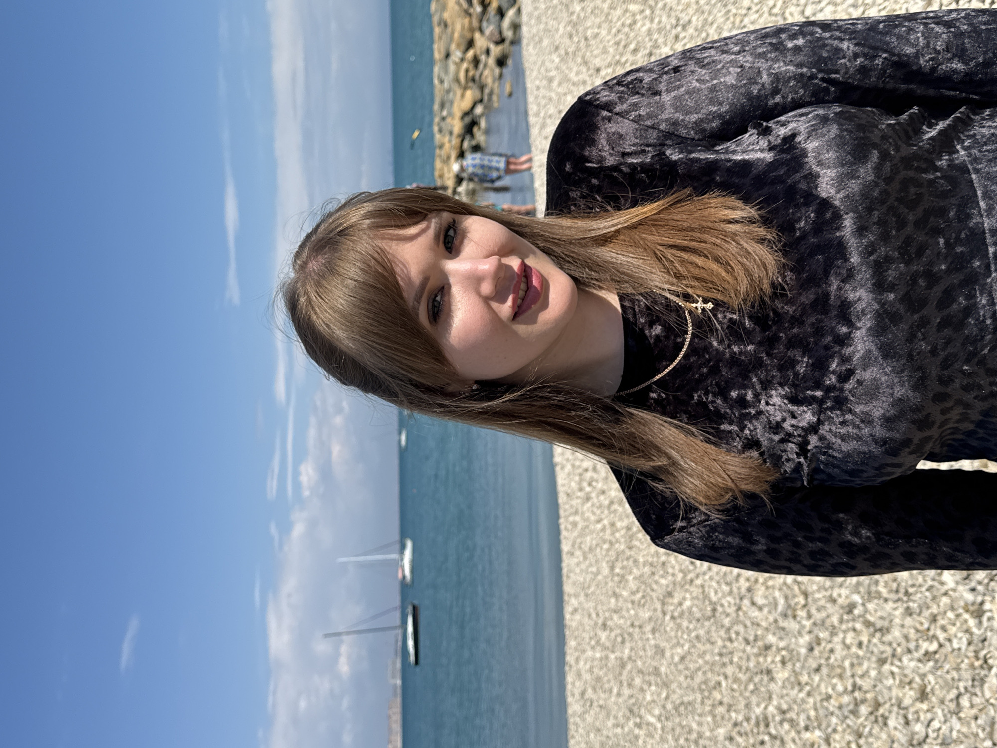 A woman poses for a photograph near the Black Sea in Yevpatoriya, Ukraine. The woman has medium-length brown hair and fair skin, wearing a dark, textured top and a gold necklace. The scene is outdoors on a clear day. In the background, the calm, blue waters of the Black Sea contain several sailboats. A stone breakwater with people standing on it is visible further in the background. The foreground is a pebble-covered ground.