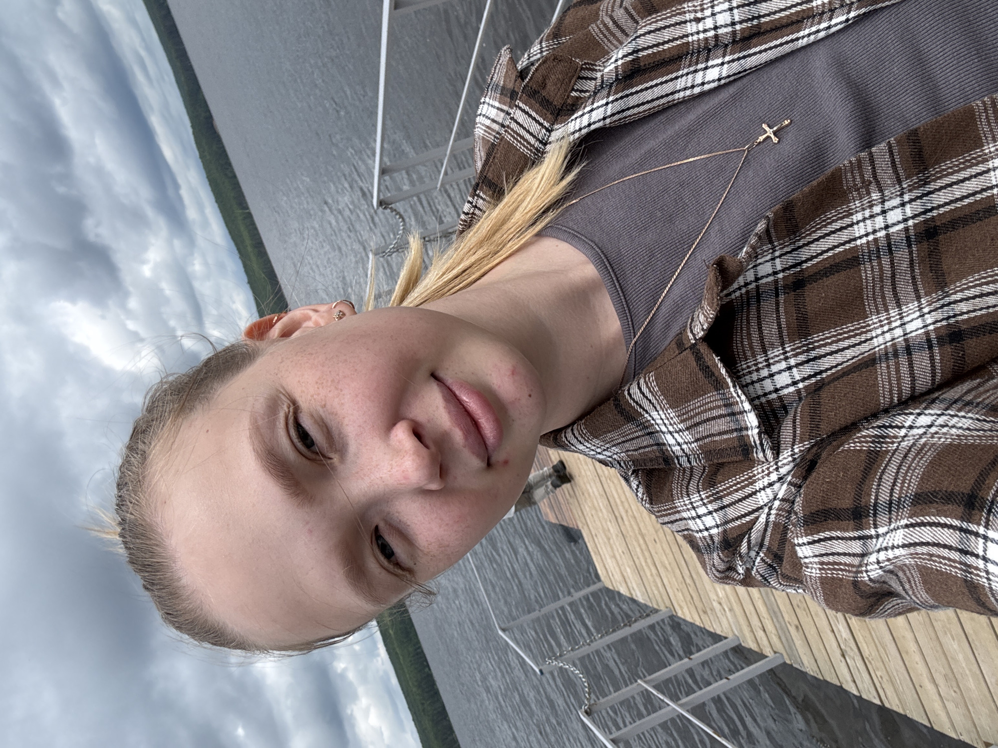 The image shows a young woman taking a selfie on a wooden pier by a body of water. She has blonde hair pulled back and is wearing a brown and white plaid shirt over a grey top. A thin necklace with a cross pendant is visible. The background features a lake or river with a tree-lined shore and a cloudy sky. The setting appears to be outdoors, likely in a natural or semi-natural environment. Without specific landmarks, it is impossible to definitively place this scene within the city of Chaikovsky, Russia, though it is consistent with the general landscape of the region, which features numerous lakes and forests.