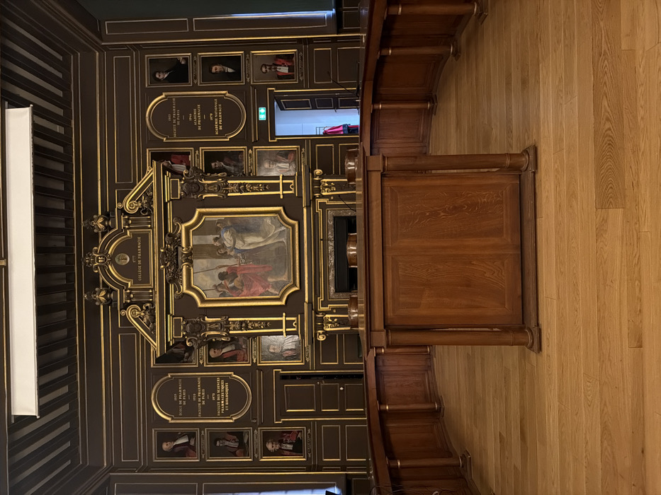 The image captures the Salle des Séances (Meeting Room) of the Académie Nationale de Pharmacie, located at 4 avenue de l'Observatoire, 75006 Paris, France.
The scene presents a formal, empty chamber dominated by dark brown wooden wall paneling extensively decorated with gold trim and ornate carvings. Centered on the main wall is a large, elaborate golden-framed oil painting depicting a historical or allegorical scene with multiple figures, primarily men in robes and period attire, clustered around a central, light-skinned individual. Below this painting is a dark fireplace mantel. Flanking the central artwork are numerous smaller framed portraits of historical figures and several gold-lettered plaques detailing the lineage of Parisian pharmacy institutions, including "COLLEGE DE PHARMACIE" (1777), "ECOLE DE PHARMACIE DE PARIS" (1803), and "ACADEMIE NATIONALE DE PHARMACIE" (1979).
In the foreground, a substantial assembly of light-colored polished wooden furniture forms a U-shaped arrangement. This includes a prominent rectangular central rostrum or desk with two paneled doors facing the viewer, flanked by a curved table section extending to the left and right, supported by decorative fluted columns. Several chairs with dark backs are visible behind the curved table. The floor is composed of wide, polished light hardwood planks.
A dark wooden door, marked by a green "exit" sign above, is partially open on the right side of the main wall, revealing a glimpse of a brightly lit room beyond, where red and black academic robes hang on a rack. The ceiling is dark wood-paneled, featuring a long, rectangular white light fixture. No individuals are present in the immediate room.