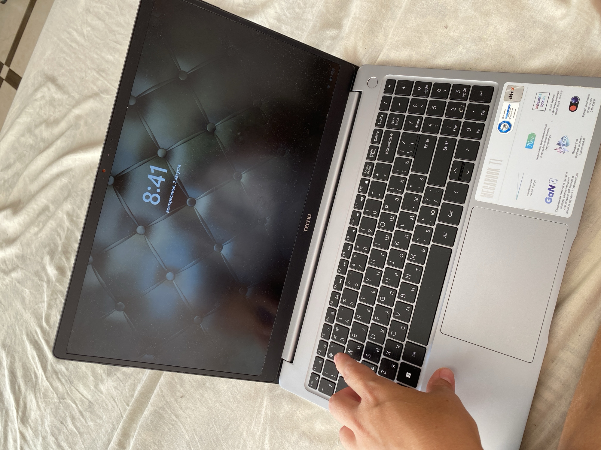 A silver laptop is open on a light-colored, textured surface, possibly a bedspread, with a checkered floor visible in the background. The laptop screen displays a time of 8:41 and the date "воскресенье, 2 августа" (Sunday, August 2nd) on a dark, quilted wallpaper. A person's hand is visible, with the index finger touching the 'Q' key on the keyboard. The laptop has a Russian keyboard layout. Stickers with information like "MEGABOOK T1", "GaN", "70Wh", "100%sRGB 350nits", and logos like "TÜV" and "dts" are present on the bottom right corner of the laptop's base. The overall scene is indoors, well-lit, and suggests a casual setting. There is no information in the image to suggest a location in Sochi, Russia.