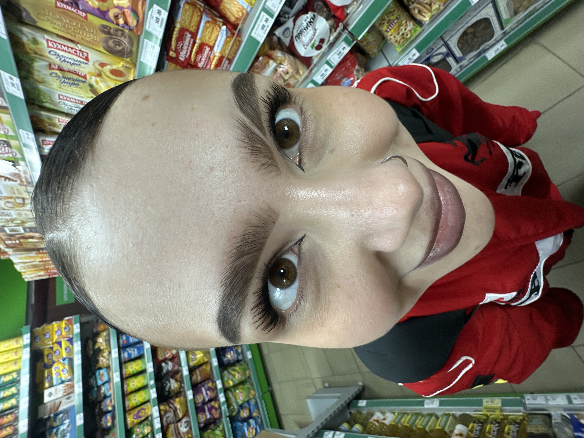 A person is pictured close-up, possibly taking a selfie, in a grocery store aisle. Shelves filled with snacks and other grocery items are visible in the background. The person has brown eyes, dark hair, and distinct eyebrows. They are wearing a red jacket with white accents over a black shirt. The setting seems to be a commercial area, likely a supermarket. Given the Cyrillic script on some of the product packaging, this is Moscow, Russia.