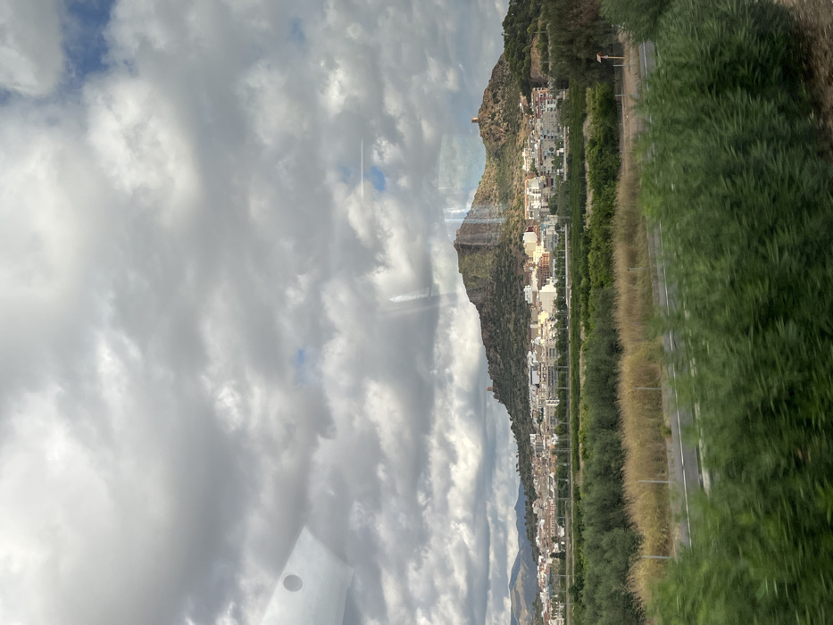 The image depicts a panoramic landscape of Almenara, Spain, observed under a cloudy sky with intermittent blue patches. The upper half of the frame is dominated by a dense formation of grey and white clouds, with a visible circular reflection in the upper left quadrant and several vertical light streaks across the frame, consistent with reflections off a window.
In the background, a series of hills and mountains rise, with the central and most prominent hill exhibiting rugged, exposed rock formations and sparse green vegetation. Situated at the base and extending partway up these hills is the city of Almenara, characterized by numerous light-colored buildings with varied rooflines. Two distinct, brown-toned tower structures, likely historical fortifications (e.g., Almenara Castle remnants), are visible on separate peaks/ridges of the main hill, one to the left of the central peak and another to the right.
The midground features cultivated land, consisting of rows of dense green trees, indicative of orchards or groves, interspersed with patches of dry, lighter-colored vegetation. A two-lane road or highway, demarcated by white lines and a thin barrier or guardrail, runs horizontally through this area.
The foreground is occupied by blurred green foliage, likely shrubs or bushes, suggesting proximity to the camera and motion blur, consistent with a photograph taken from a moving vehicle. No individuals or specific human activities are discernable within the scene.