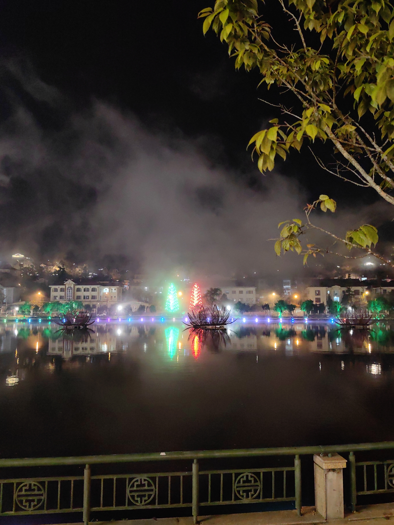 A nighttime scene depicts Sa Pa Lake in Sa Pa, Vietnam, under heavy fog or low clouds. The foreground shows a dark green metal railing with recurring traditional "Shou" (longevity) symbols, supported by a light-colored concrete post on the right. The lake occupies the midground, its dark surface reflecting numerous artificial lights from the background. Floating on the lake are five illuminated decorative structures: three large, dark-colored lotus flower-shaped sculptures with internal lighting (one exhibiting blue, one green, one red/orange hues), and two conical, tree-like structures (one green, one red/orange). A string of multi-colored lights lines the far bank of the lake. In the background, buildings of varying sizes and styles, including a prominent white-facaded structure with arched windows, are illuminated, their lights piercing through the dense fog that obscures the upper portions of the city and the distant hillsides. A tree branch with green foliage extends into the frame from the top right. No individuals are present.