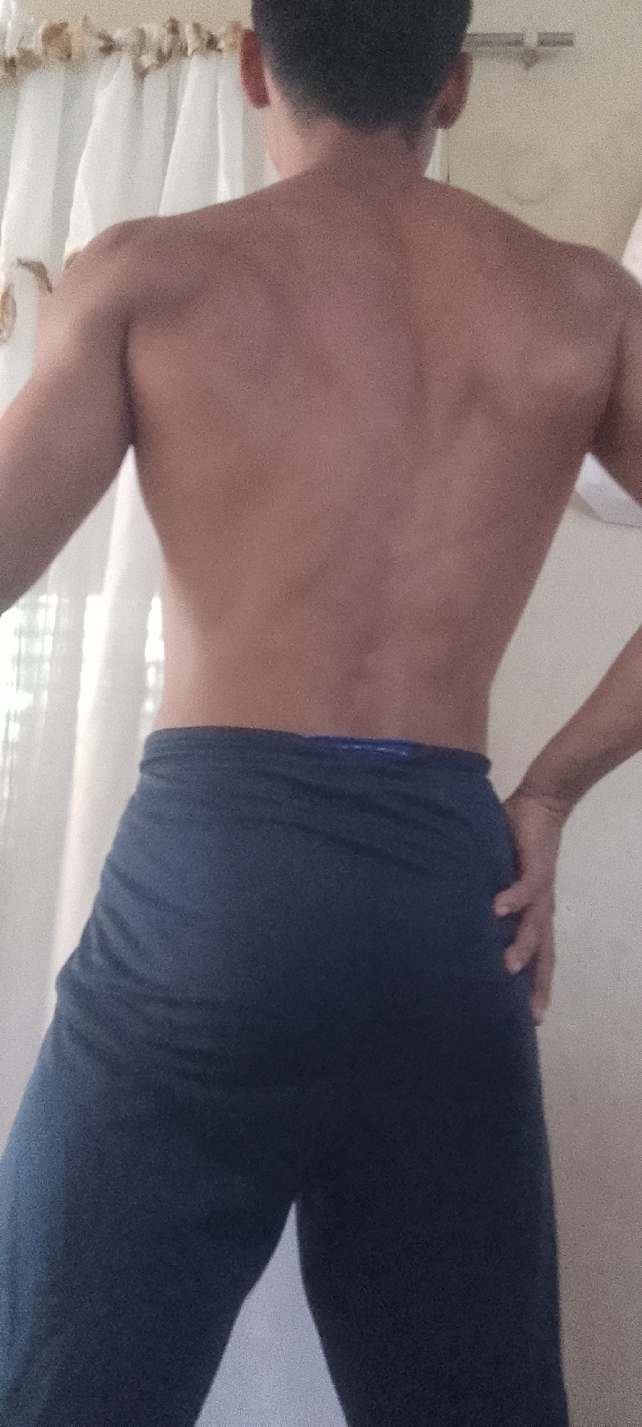 A shirtless person is shown from the back, wearing dark blue pants. The person's right hand is placed on their right hip. A curtain is visible in the background. The location in Juan Griego, Venezuela is not discernible from the image.