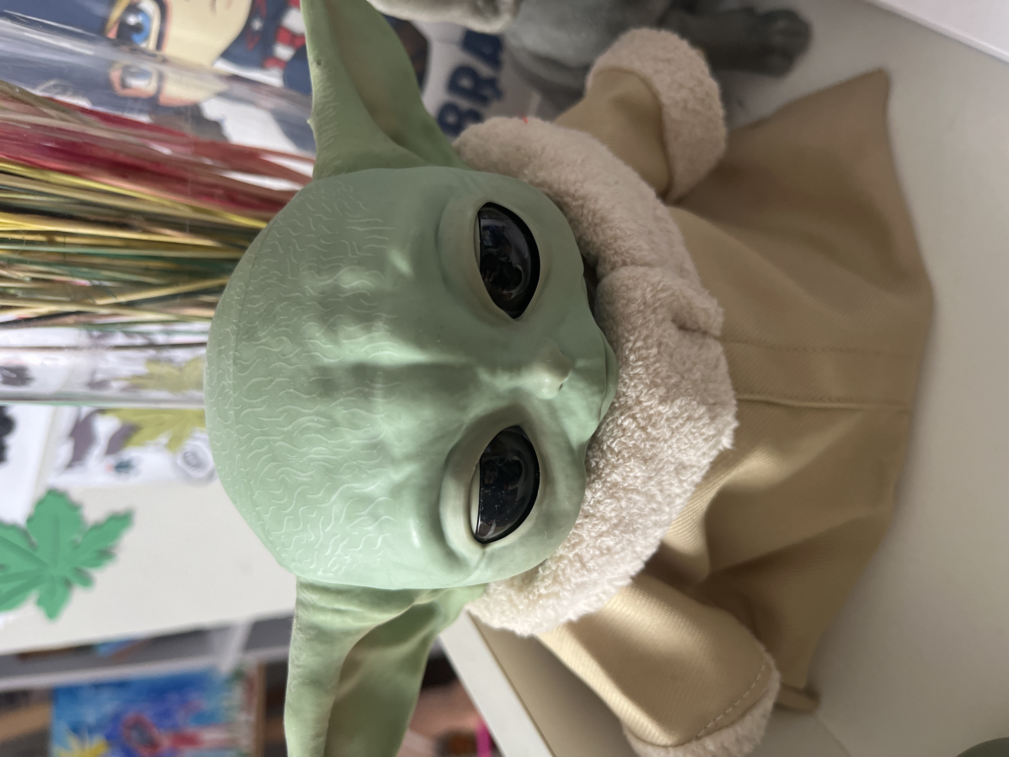 A close-up, eye-level shot captures a light green, doll-like figurine resembling a character from Star Wars, specifically Grogu. The figurine has large, black eyes and is dressed in a tan robe with a fluffy white trim. The background is blurred, showing various objects including colorful sticks in a container, a painting with a blue and yellow abstract design, and what appears to be an anime-style drawing of a person with blue eyes and glasses. The figurine is positioned in the foreground, resting within a white box or shelf. The location is not specified in the image or hints, therefore it cannot be provided.