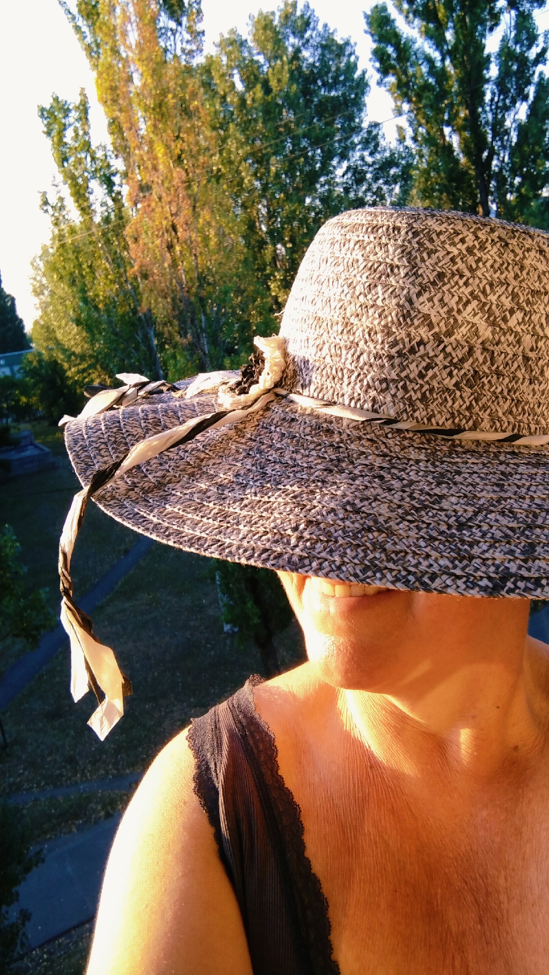 The image is a close-up, outdoor shot of a person wearing a wide-brimmed, textured hat. The hat is decorated with a ribbon and a flower. The person's face is partially obscured by the hat, revealing their mouth and chin. The person is also wearing a black, lace-trimmed garment. The background consists of trees and a hint of what appears to be a park or residential area. The lighting suggests it is either early morning or late afternoon, with warm sunlight illuminating the person's skin. This image was captured in Horishni Plavni, Ukraine.