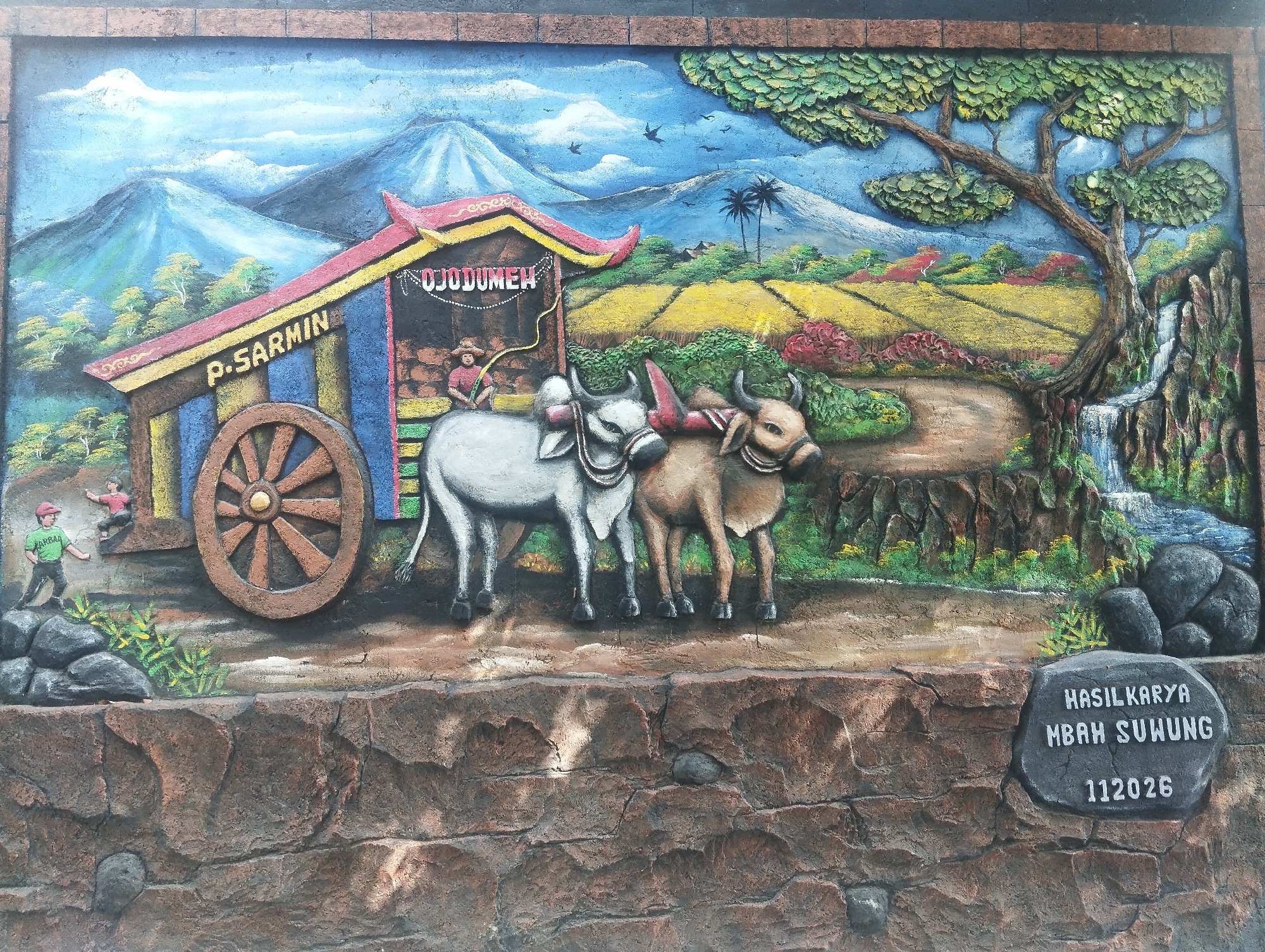 This is a detailed, three-dimensional mural depicting a rural Indonesian scene. In the foreground, two oxen are yoked together, standing in front of a wooden cart. The cart, labeled "P. SARMIN" and "OJODUMEN," has a colorful, traditional roof and a large wooden wheel. A figure in a straw hat appears to be operating the cart. To the left, two small figures of people, possibly children, are depicted, one near the cart and another in the distance.
The background of the mural showcases a vibrant landscape with rolling hills, lush green trees, and fields of yellow and red crops. Majestic blue mountains with snow-capped peaks rise in the distance under a bright blue sky with scattered clouds and birds in flight. To the right, a waterfall cascades down a rocky cliff into a stream, surrounded by dense foliage. A large, gnarled tree with green leaves dominates the right side of the scene, its branches reaching towards the sky.
At the bottom right of the mural, a stone-shaped plaque is integrated into the artwork. It bears the inscription "HASILKARYA MBAH SUWUNG" followed by the number "112026."
This mural is located on the exterior wall of a building in Malang, Indonesia, likely as a decorative piece or a form of public art.