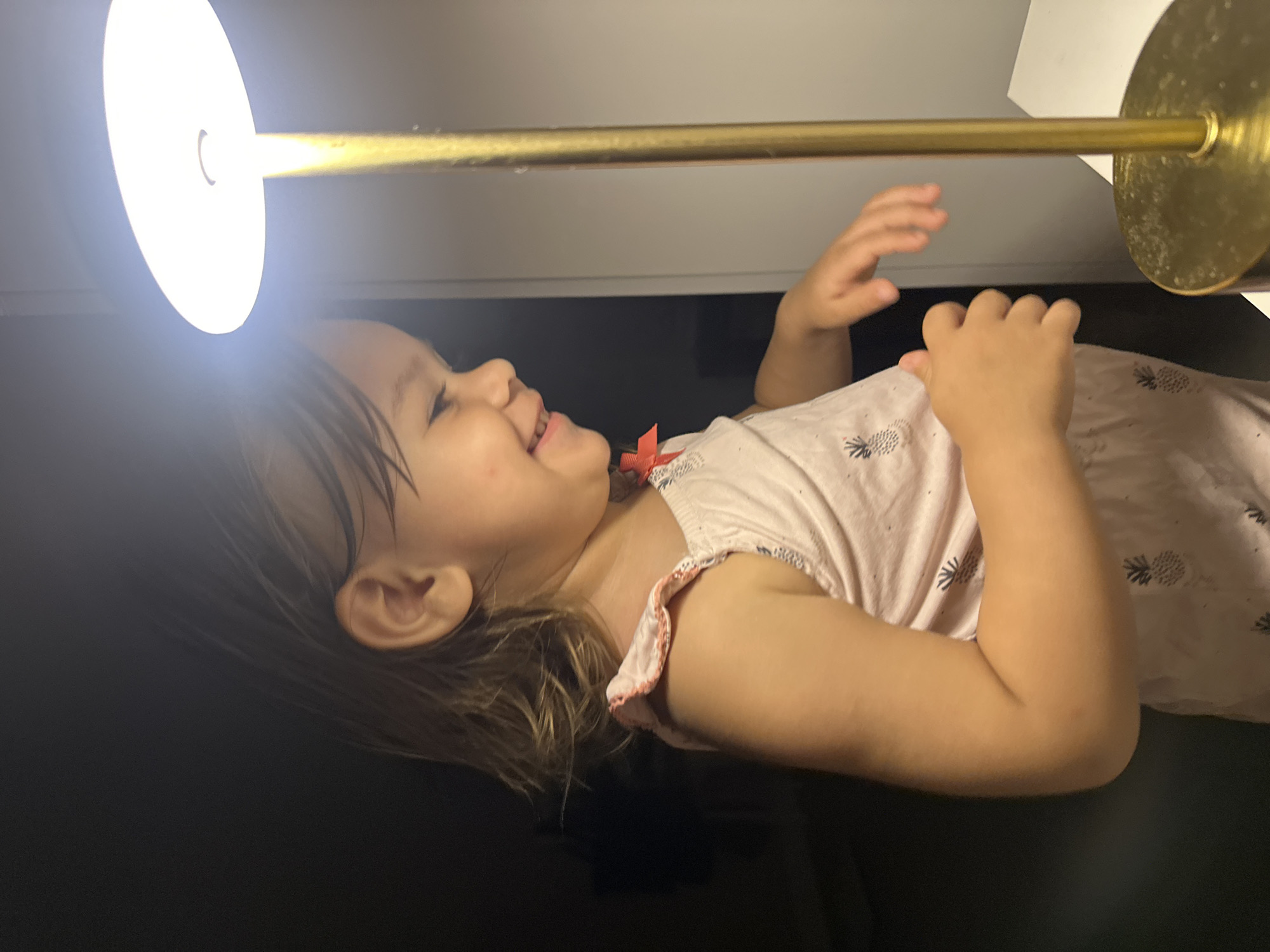 A young child, appearing to be between two and four years old, is observed in a dimly lit indoor environment, possibly a bedroom or living area. The child is positioned in profile, facing towards the right side of the frame, and is illuminated by the bright light of a brass table lamp. The child has light brown hair, parted on the left and reaching just below the ears. They are wearing a light pink sleeveless top adorned with a repeating pattern of pineapples, and the top has ruffled shoulder straps with a pink trim. A small red ribbon is visible on the left shoulder strap. The child's facial expression is one of mirth and curiosity, with an open mouth and what appears to be a smile. Their right hand is extended slightly forward, while their left arm is bent at the elbow, with the hand near the chest. The background is predominantly dark, with the exception of a light-colored surface on the right, likely a table or shelf, where the lamp is placed. The lamp itself has a round, white lampshade and a thin brass stem and base. There are no discernible features indicating the specific location within Kharnavy, Russia, as the environment is generic and lacks identifiable landmarks or architectural details.
