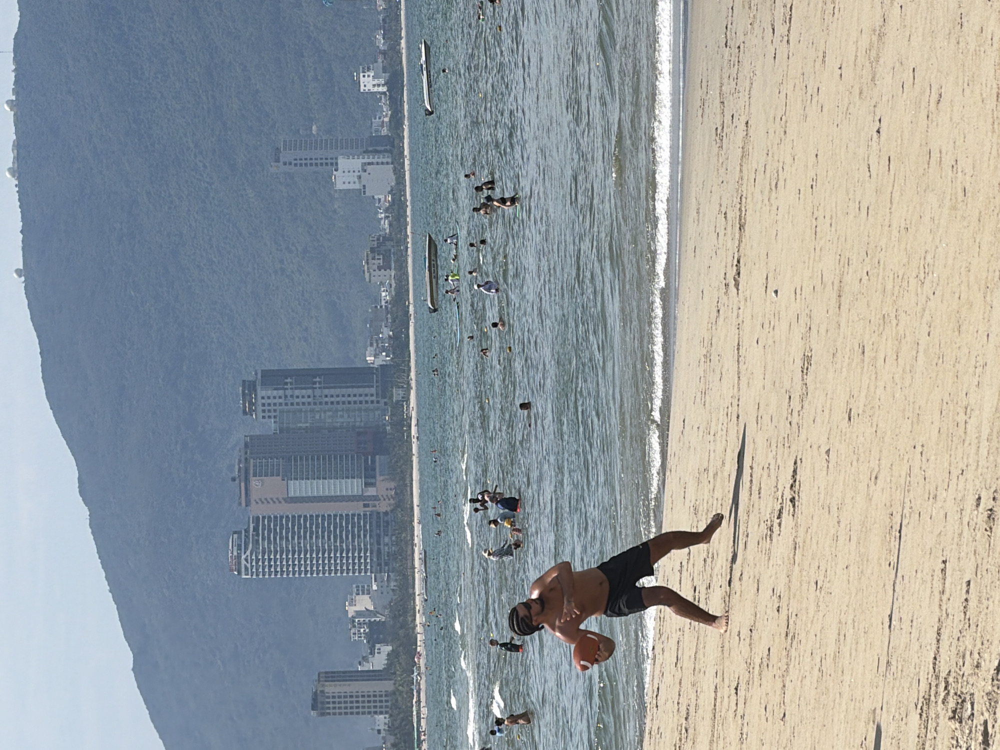In Đà Nẵng, Vietnam, a man stands on a sandy beach, holding an orange football. He is shirtless, wearing black shorts, and appears to be in the process of throwing the ball. In the background, the ocean is filled with people swimming and a few boats are visible. Beyond the water, a city skyline with tall buildings rises, backed by a large, tree-covered mountain. The sky is bright and clear.