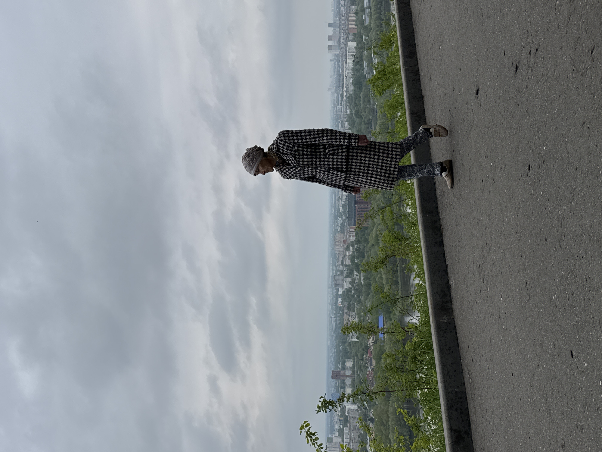 A person is standing on a paved area with a city visible in the background, most likely in Nizhny Novgorod, Russia. The person, wearing a patterned coat and head covering, is the main subject. The city sprawls in the distance, interspersed with greenery. The sky is overcast. The paved area extends along the right edge of the frame. Trees are growing alongside the pavement.