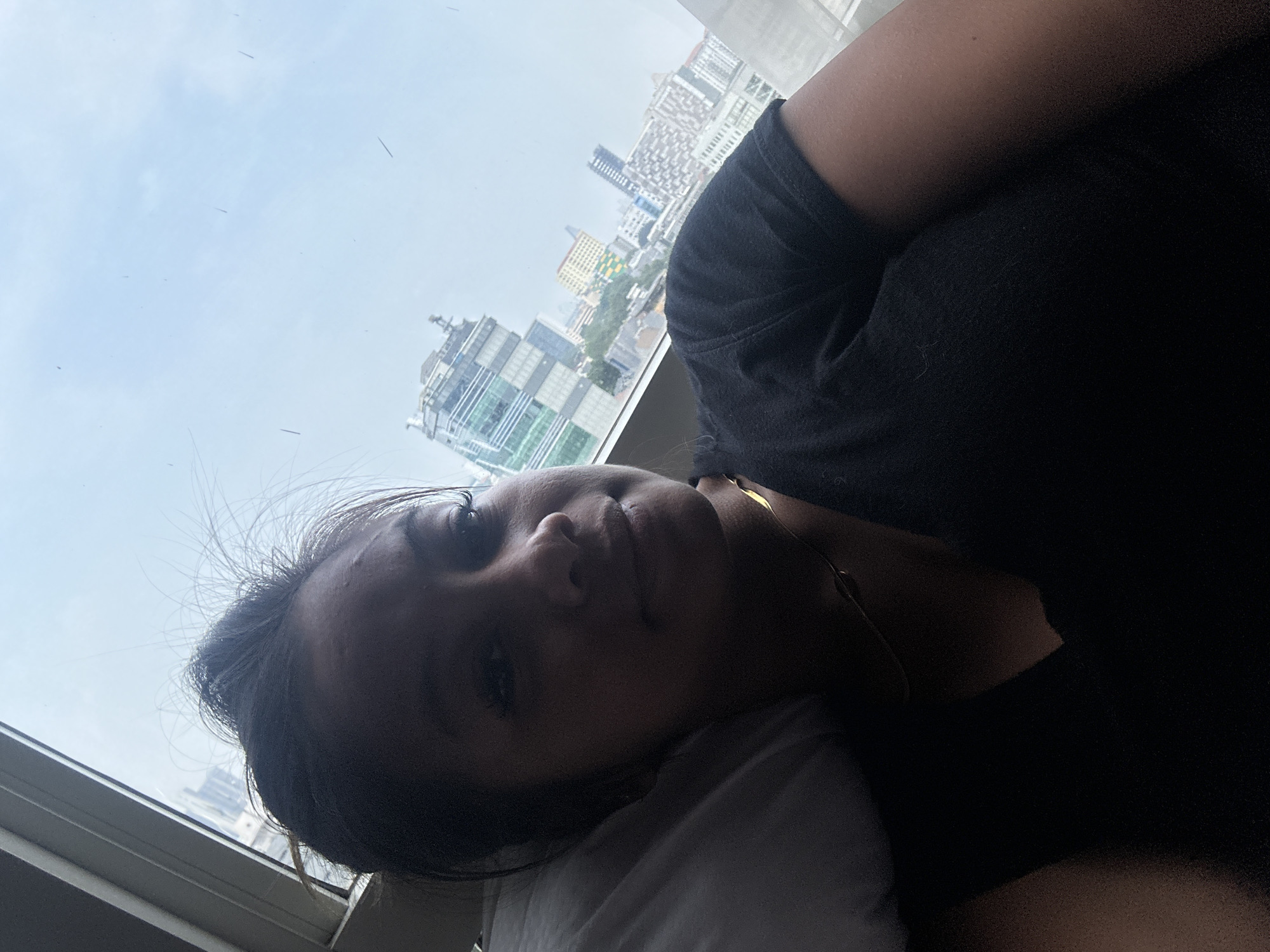 A woman is lying down in what appears to be a room with a large window. The window provides a view of a cityscape, with several tall buildings visible under a cloudy sky. The buildings are characteristic of a dense urban environment. Based on the architecture and general skyline, the location is likely in the city of Central Jakarta, Indonesia. The woman is wearing a dark-colored shirt and is resting her head on a white pillow.