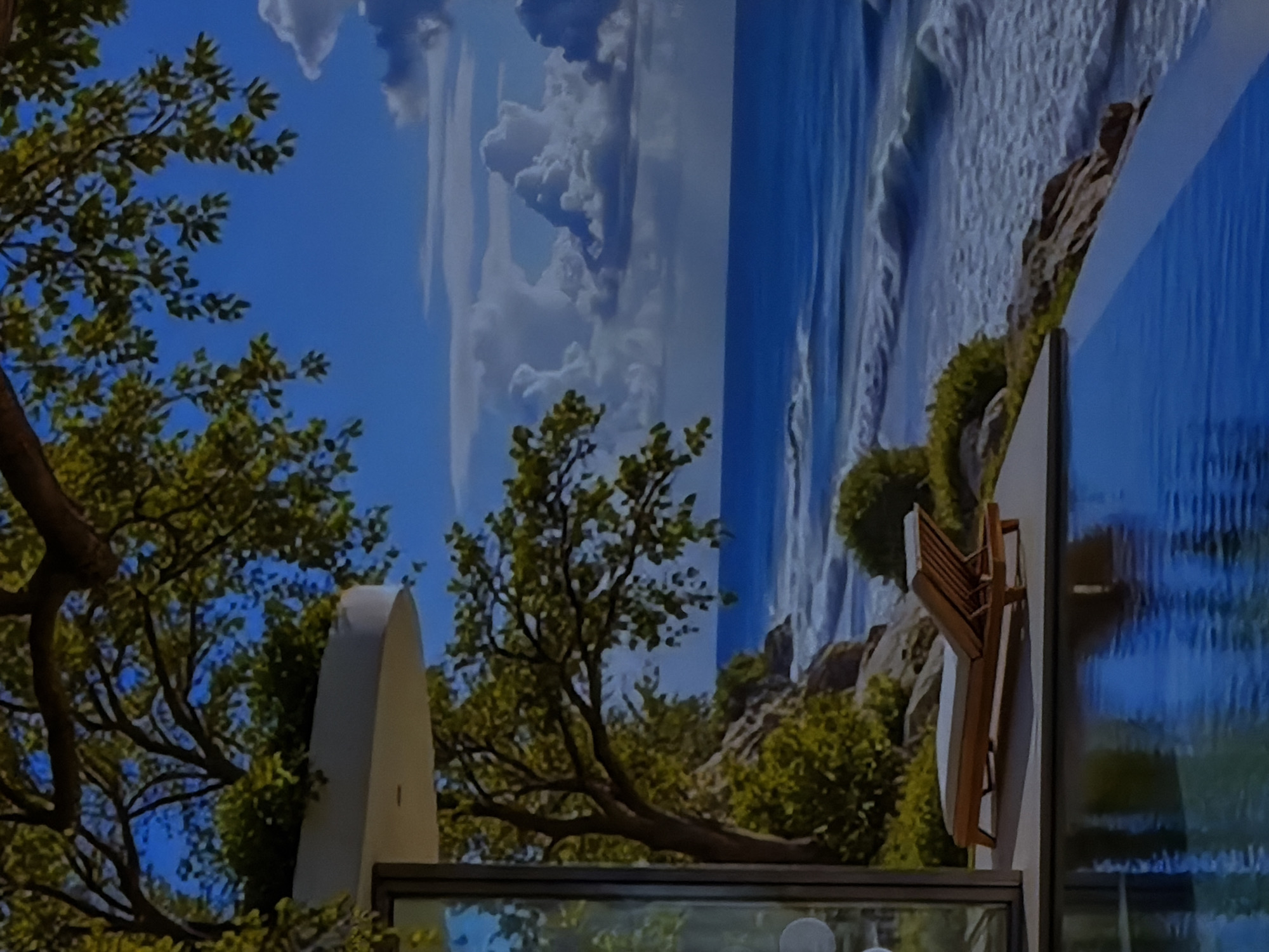 The image displays an interior space that skillfully integrates a large, vibrant digital display or mural depicting a natural landscape with real-world foreground elements. The main background is dominated by this expansive display, which shows a brilliant blue sky dotted with soft, white cumulus clouds, indicative of a clear daytime setting. On the right side of the digital scene, a dramatic natural feature unfolds: a wide expanse of water, either a powerful ocean with white-capped waves crashing or a majestic waterfall cascading down, its deep blue hues contrasting with the foamy white. The digital landscape also features lush green trees and rocky formations, creating a rich, natural environment.
In the immediate foreground, real or highly realistic artificial trees with dark branches and vibrant green leaves extend into the frame from the left and bottom-center, seemingly blending with the displayed scenery. A smooth, white, arch-like architectural element is partially visible on the bottom-left, nestled among the foliage. On the right side, an intriguing detail is a wooden chair, mounted vertically on a wall, appearing to be folded flat or designed to hang. Adjacent to it, a white, circular table surface is also oriented vertically, suggesting a space-saving design or an artistic installation. Below these items, a horizontal, reflective surface, likely glass or a polished counter, mirrors parts of the vibrant display above.
There are no people present, and no specific activity is taking place, creating a serene and contemplative atmosphere. The scene evokes a feeling of being immersed in nature despite being indoors, suggesting a modern design choice for a commercial or residential space in Sofiivska Borshchahivka, Ukraine. The visual elements imply a sunny, pleasant day. No visible text is present in the image.
