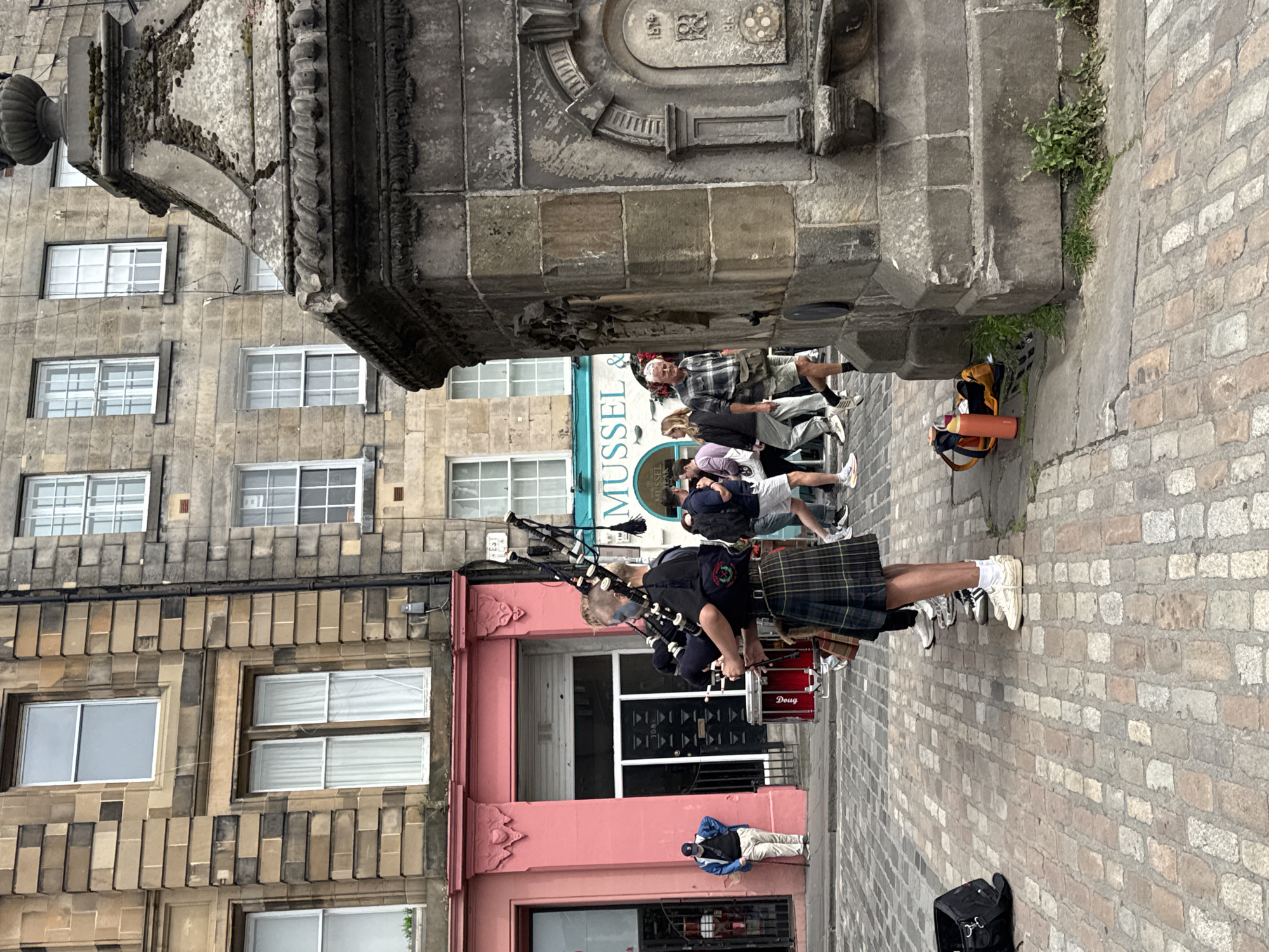 The image shows a street scene in the City of Edinburgh, United Kingdom. A bagpiper dressed in traditional Scottish attire, including a kilt, is playing bagpipes in the foreground. A red drum is positioned next to the bagpiper. Several people are walking in the background. Buildings with stone facades and windows line the street. A stone fountain is visible on the right side of the image. The ground is paved with cobblestones.