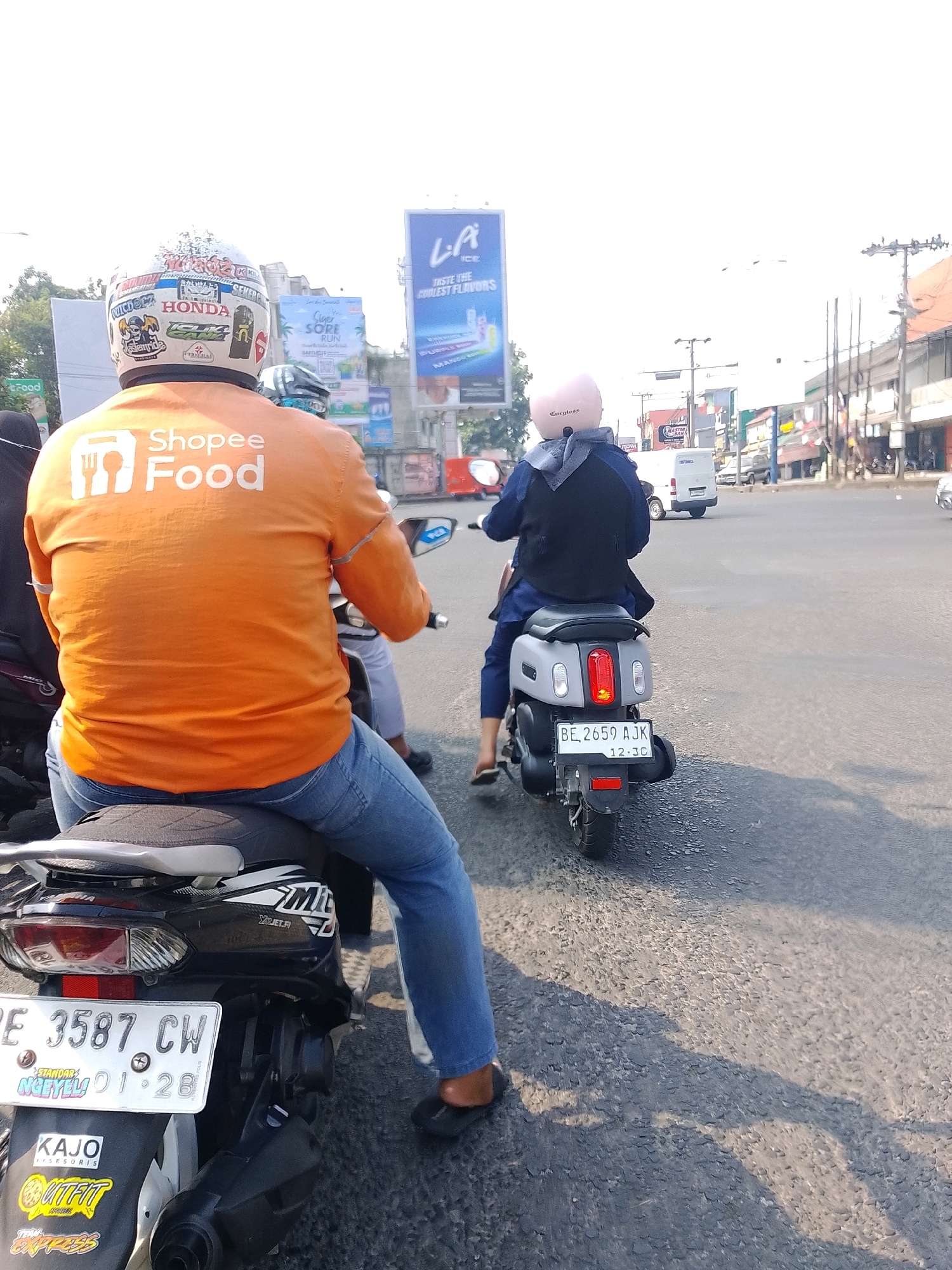 The image shows a street scene in Bandar Lampung, Indonesia, on a sunny day. In the foreground, a person wearing an orange Shopee Food jacket and blue jeans is sitting on a black motorcycle. The motorcycle has a license plate that reads "BE 3587 CW" and several stickers, including "KAJO AKSESORIS" and "OUTFIT APPAREL." The person is wearing a white helmet with various stickers and designs.
To the right of the foreground motorcycle, another person is riding a grey electric scooter. This person is wearing a blue jacket, a grey headscarf, and a pink helmet. The scooter has a license plate that reads "BE 2659 AJK 12.30."
In the background, there are buildings, billboards, and utility poles lining the street. One billboard displays "L.A. ICE" and advertises flavors. Another billboard has text that is difficult to read but appears to be an advertisement. Traffic lights are visible, and other vehicles, including a white van, are in the distance. The road surface is asphalt, and shadows from the vehicles and overhead objects are cast on the road. The overall environment suggests a typical urban street in Indonesia during daylight hours.