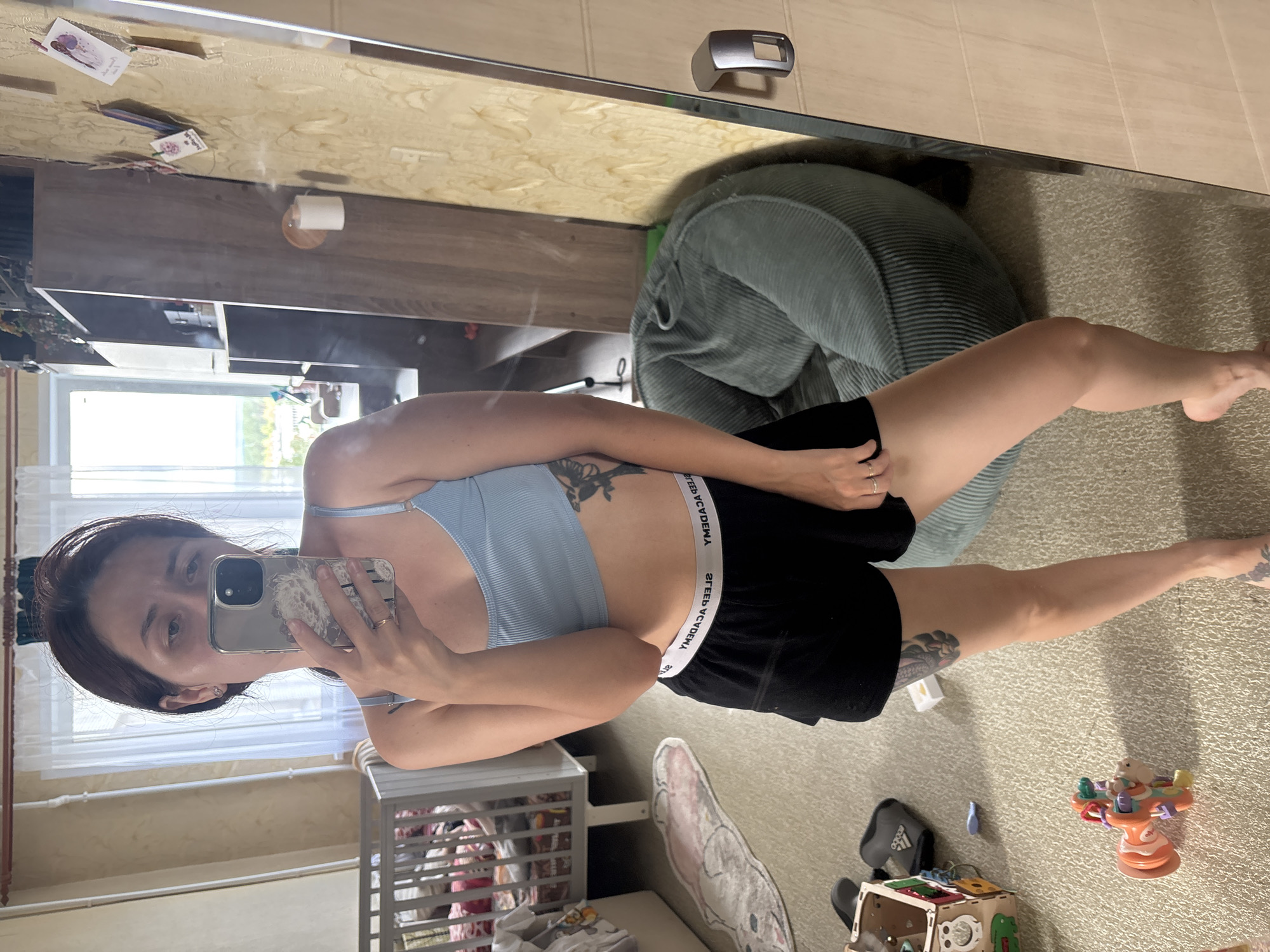 A woman is taking a mirror selfie in a room. She is wearing a light blue ribbed tank top and black shorts with a white waistband that reads "SLEEP ACADEMY." She has dark brown hair pulled back in a ponytail and is wearing a ring on her left hand. There are tattoos visible on her arm and leg. The room appears to be a bedroom or nursery, with a crib visible in the background. A large beanbag chair is to her right, and a mirror is to her right, reflecting more of the room. The floor is carpeted and there are various toys and objects scattered around the room. The location is identified as Naberezhnye Chelny, Russia.
