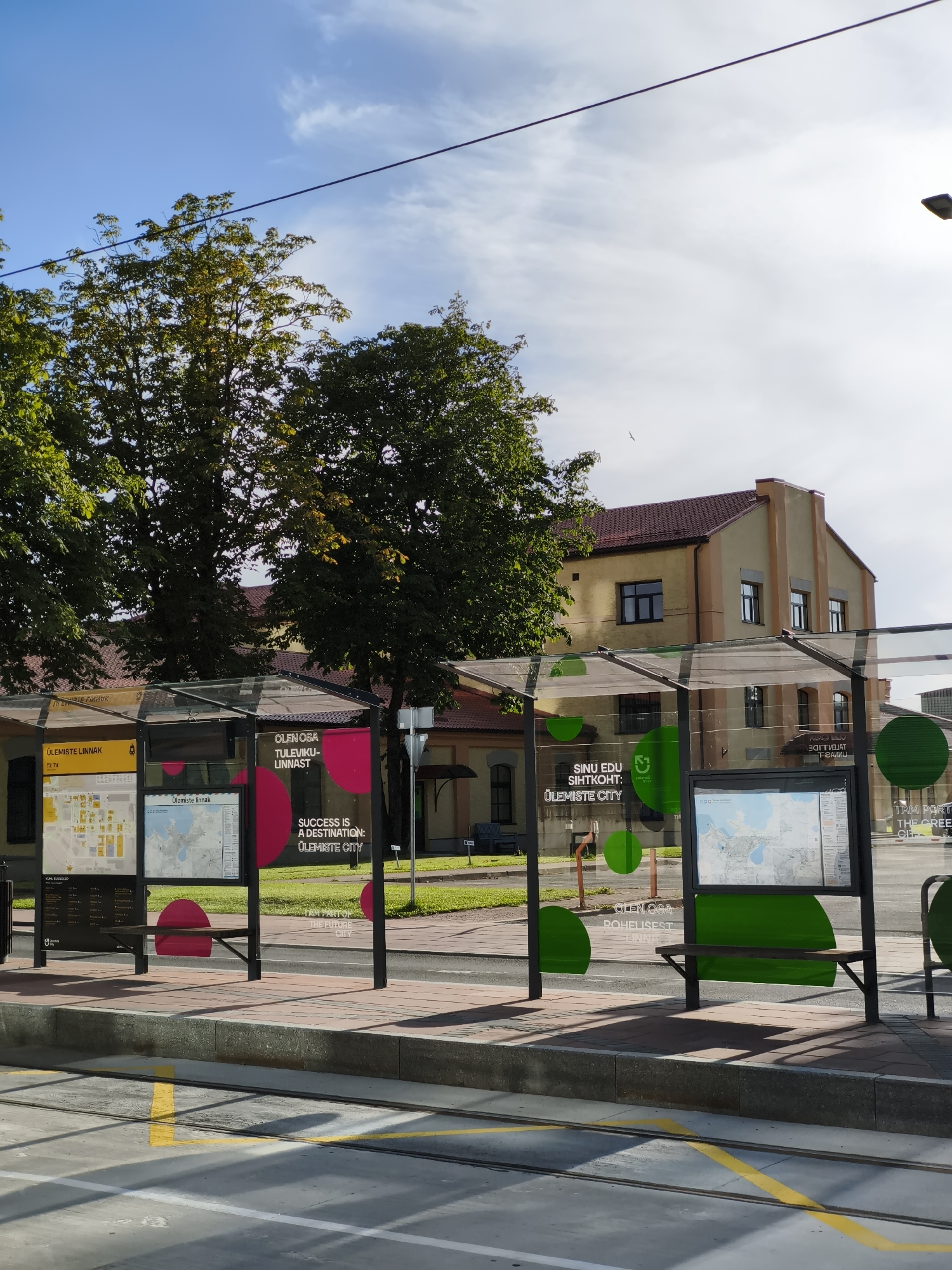 This image captures a sunny day scene at the Ülemiste City tram stop in Tallinn, Estonia. The foreground features tram tracks with yellow and white markings. The tram stop itself has a modern, glass-enclosed design with a sloped roof supported by black metal poles. Inside the shelter, there are informational displays with maps and text, along with dark green benches for seating. Advertisements are visible on the glass panels. The background reveals a building with a reddish-brown roof and light yellow walls, featuring multiple windows. Lush green trees with some autumnal foliage frame the scene, suggesting it's either late summer or early autumn. The sky is predominantly blue with scattered white clouds. No people are visible in the frame.