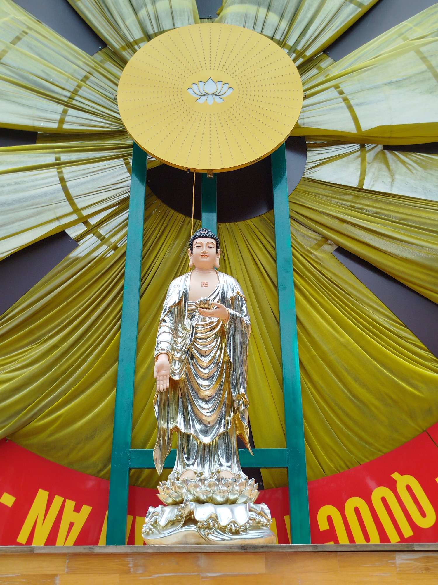 A polished silver statue of Buddha stands on a lotus base. The statue is centered, framed by a green metal structure supporting a yellow disc with a lotus emblem. A yellow draped backdrop fills the space behind the statue. The scene is set on a wooden platform with a red banner featuring yellow text, partially obscured. The location is Liên Nghĩa, Vietnam.