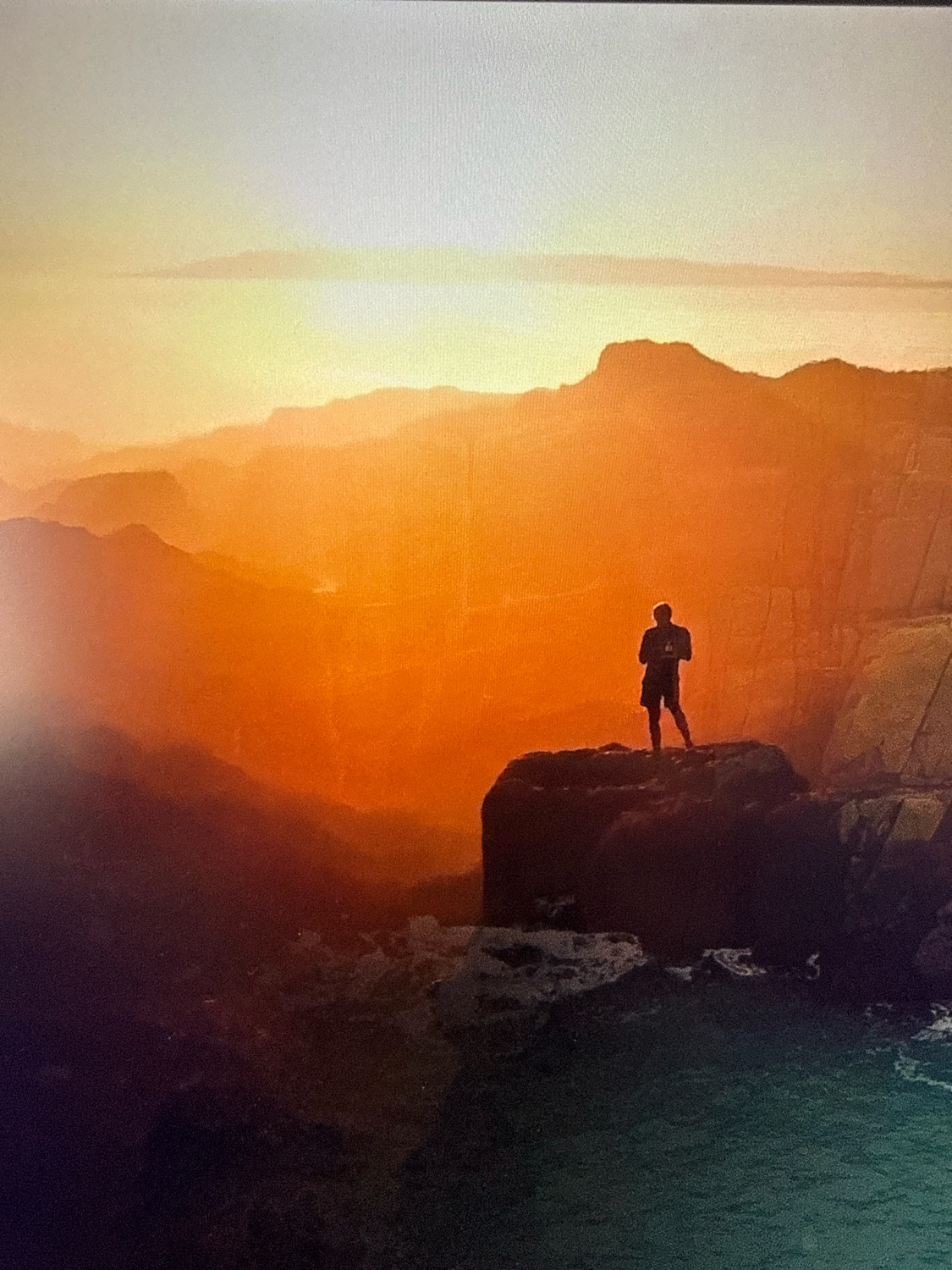The image depicts a solitary figure standing on a rocky outcrop overlooking a vast expanse of mountains at sunset. The sky is a gradient of warm oranges and yellows, transitioning to a pale blue at the top. Layered mountain ranges stretch into the distance, their peaks softened by the atmospheric haze. The foreground features dark, rugged rocks and the suggestion of water below. The figure is silhouetted against the bright sky, wearing shorts and a t-shirt, with their arms at their sides. The overall scene evokes a sense of awe and isolation in a natural landscape. The location is identified as the cliffs of Netanya, Israel, a city known for its coastal scenery and dramatic sea cliffs.