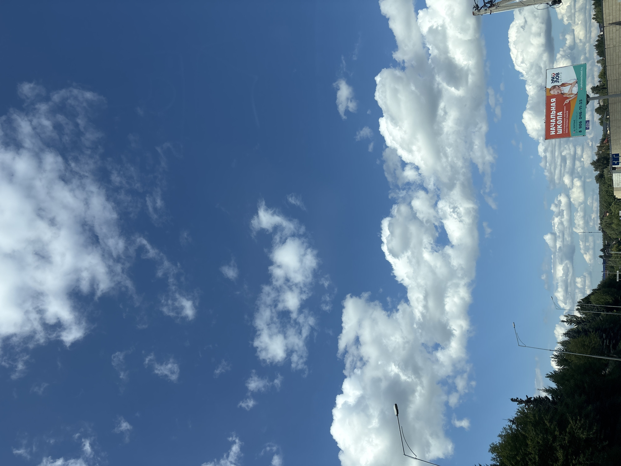 The image, taken in Obushkovo, Russia, captures a daytime scene with a vast expanse of blue sky dotted with fluffy white clouds. In the foreground, a line of dark green trees borders a road, with several streetlights visible. To the right, a billboard displays an advertisement for "НАЧАЛЬНАЯ ШКОЛА" (Primary School) featuring a young girl and contact information. The overall environment suggests a suburban or rural setting under a clear, sunny sky.