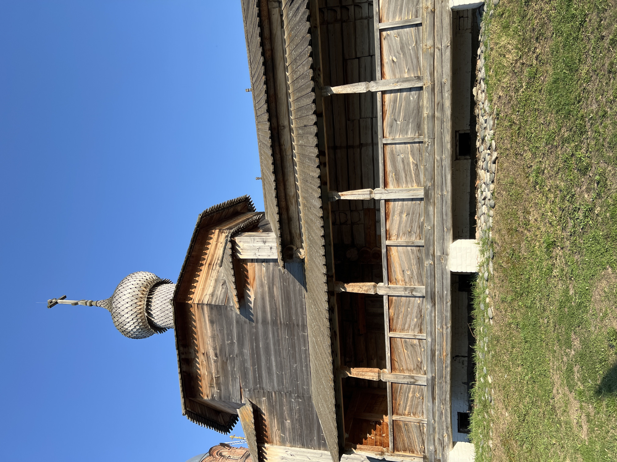 The image depicts a wooden church structure in Sviyazhsk, Russia. The structure is made entirely of wood, with a distinctive onion-shaped dome topped by a cross. The building features a tiered design with intricate wooden detailing on the roof and walls. The building appears to be old and weathered. The sky is clear and blue, and there is a patch of grass visible at the base of the building.