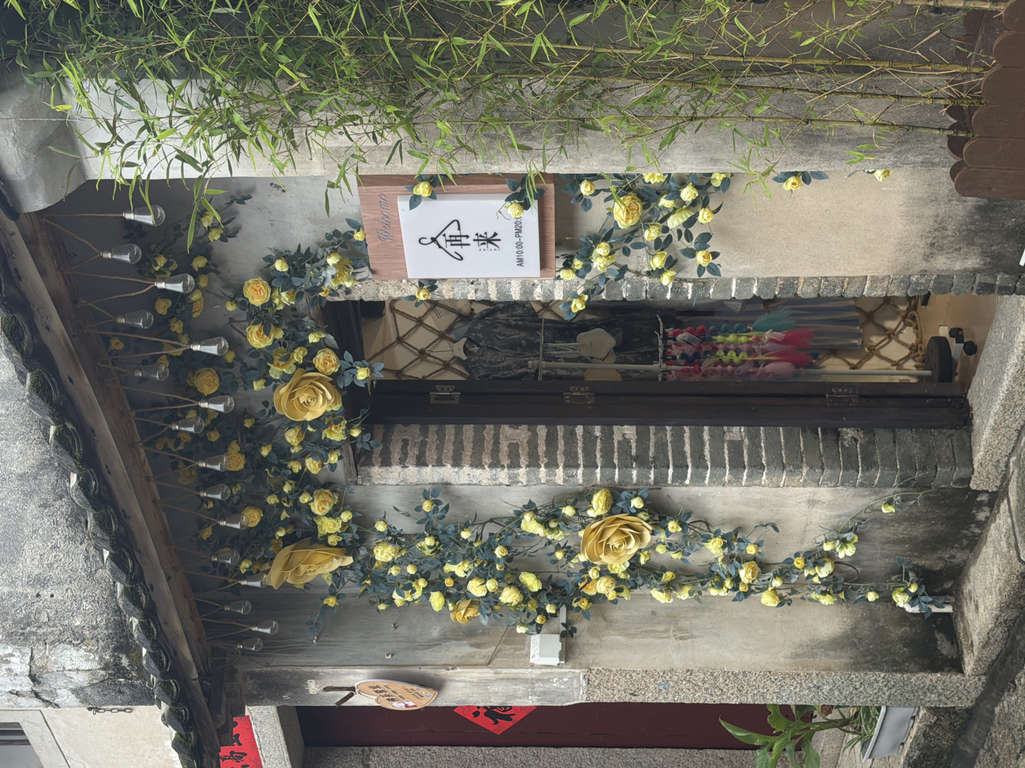 The image depicts the exterior of a shop in Longgang District, China, adorned with a profusion of yellow roses and faux greenery. The shop entrance is framed by a decorative arch of these floral elements. Hanging above the entrance are several bare lightbulbs strung on twine, adding a whimsical touch. A wooden sign with "Welcome" and Chinese characters is mounted on the wall to the right of the entrance, alongside a vertical arrangement of bamboo plants. Inside the open doorway, clothing items are visible on display racks. To the left of the entrance, a red banner with Chinese characters is affixed to a darker door. The building's facade is constructed of textured concrete and brick. The overall scene suggests a charming and inviting boutique, possibly a clothing store or a craft shop, set in an urban environment.