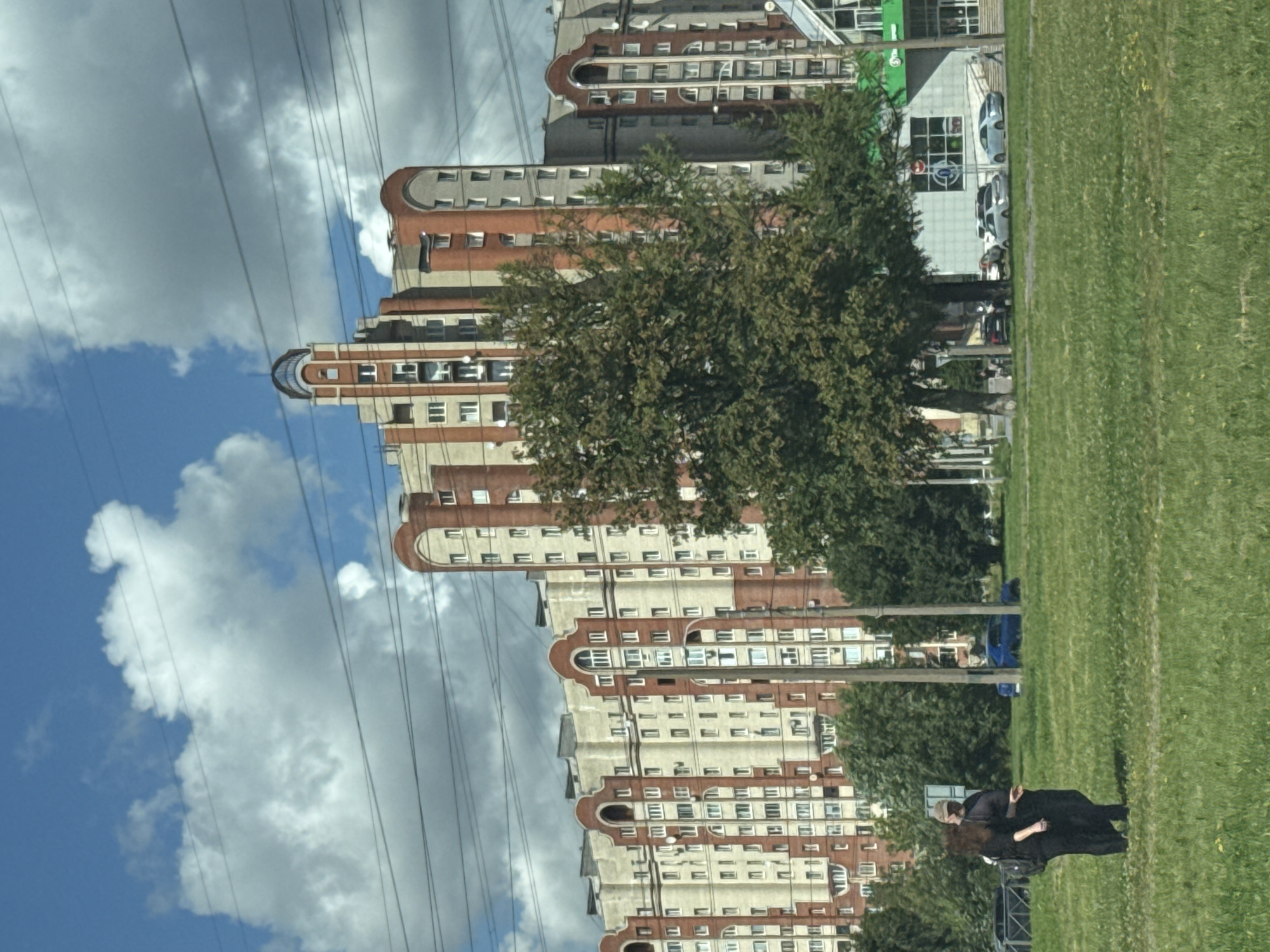 A long, multi-story apartment building with cream and red brick facade is seen under a bright blue sky with scattered white clouds. The building features arched window frames and a distinctive rounded tower on the right side. Several trees, some with green foliage, are in front of the building. A grassy lawn occupies the foreground, with two people in dark clothing walking on it. In the background, several cars are visible on a street and in a parking lot, along with a portion of a store with a green sign. Overhead, power lines crisscross the sky. This scene is likely in Saint Petersburg, Russia, given the architectural style of the apartment building.