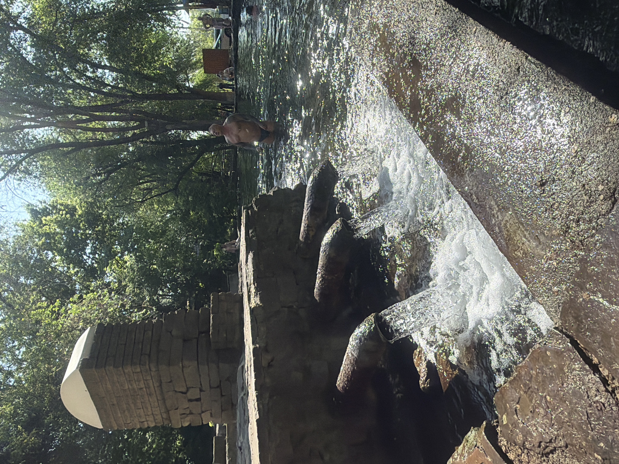 The image depicts a flowing body of water, likely a stream or river, in Rostov-on-Don, Russia. A small artificial waterfall feature is visible in the foreground, constructed of stone and concrete. Water cascades over several pipes protruding from the structure. A person is wading in the water further upstream, toward the center of the frame. Others can be seen standing near a lifeguard stand and vehicle further in the distance. The banks of the water feature are lined with trees. The lighting is bright and sunny.