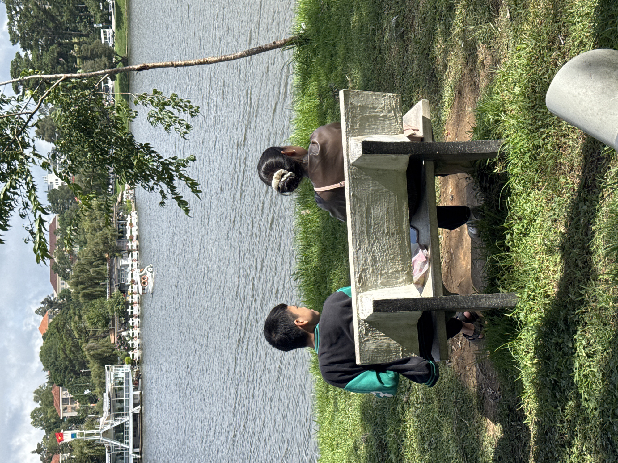 Two individuals, a young boy and an adult female, are seated on a concrete bench overlooking a lake. The boy is on the left, facing away from the camera and towards the lake. He is wearing a black long-sleeved shirt with green accents on the sleeves and collar. The adult female is on the right, also facing away from the camera. She has her hair pulled up in a bun with a scrunchie and is wearing a dark-colored top with a pink strap visible across her back. They appear to be in a park setting with lush green grass and a tree with visible branches in the foreground. In the background, across the lake, is a scenic view of buildings with red-tiled roofs, a white building with a flag, and a row of paddle boats docked along the shore. The sky is overcast. The scene is likely located in Phường Xuân Hương - Đà Lạt, Vietnam, given the presence of the lake, paddle boats, and architecture commonly found in that area.