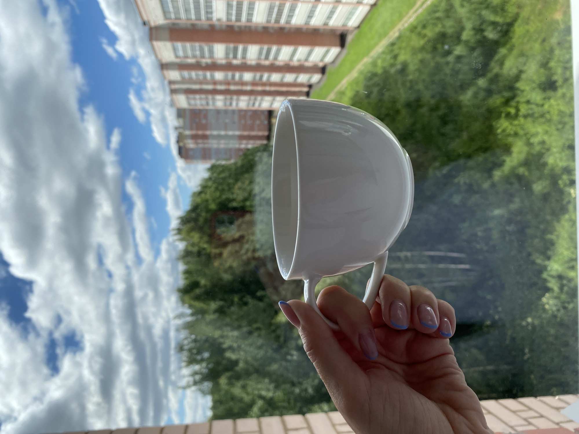 A woman's hand, with light-colored skin and manicured fingernails adorned with blue tips, holds a white ceramic coffee cup with a handle. The cup is positioned in the foreground, in front of a window. Through the window, a green, treed hillside slopes down towards a residential area. In the background, several tall, multi-story apartment buildings with light-colored facades and orange brick accents are visible. The sky above is a bright blue, dotted with fluffy white clouds. The scene appears to be a view from an apartment window, likely in a suburban or city outskirts area of Cherepovets, Russia, given the architectural style of the buildings.