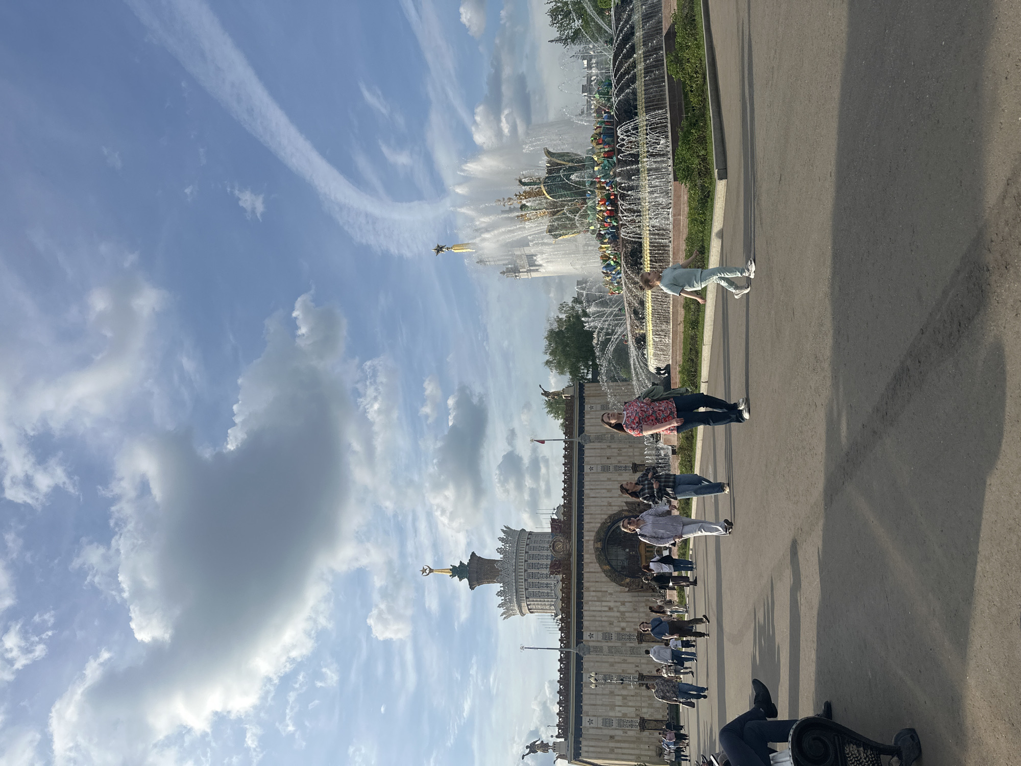 The image shows a view of the Friendship of Nations fountain and the central pavilion at the Exhibition of Achievements of National Economy (VDNKh) in Moscow, Russia. The fountain is in the foreground, with water spraying high into the air. The central pavilion stands behind the fountain, showing its ornate architecture and golden spire topped with a star. People are walking along the pavement in front of the pavilion. The sky is blue with scattered clouds.