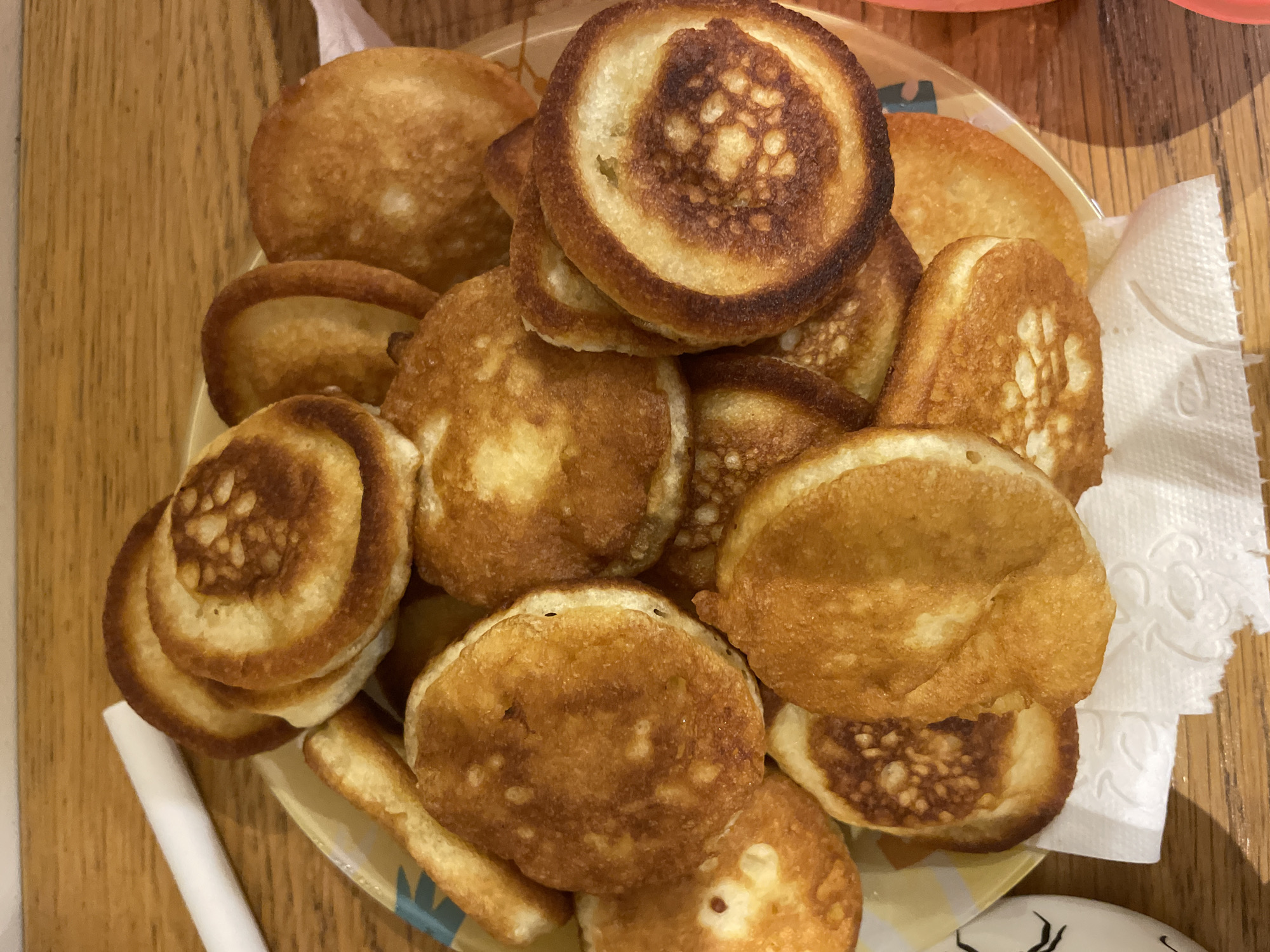 The image depicts a stack of golden-brown pancakes on a yellow plate with blue designs, resting on a wooden surface. The pancakes vary in size and color, some having darker edges. A white napkin is partially visible to the right of the plate. The location is unspecified.