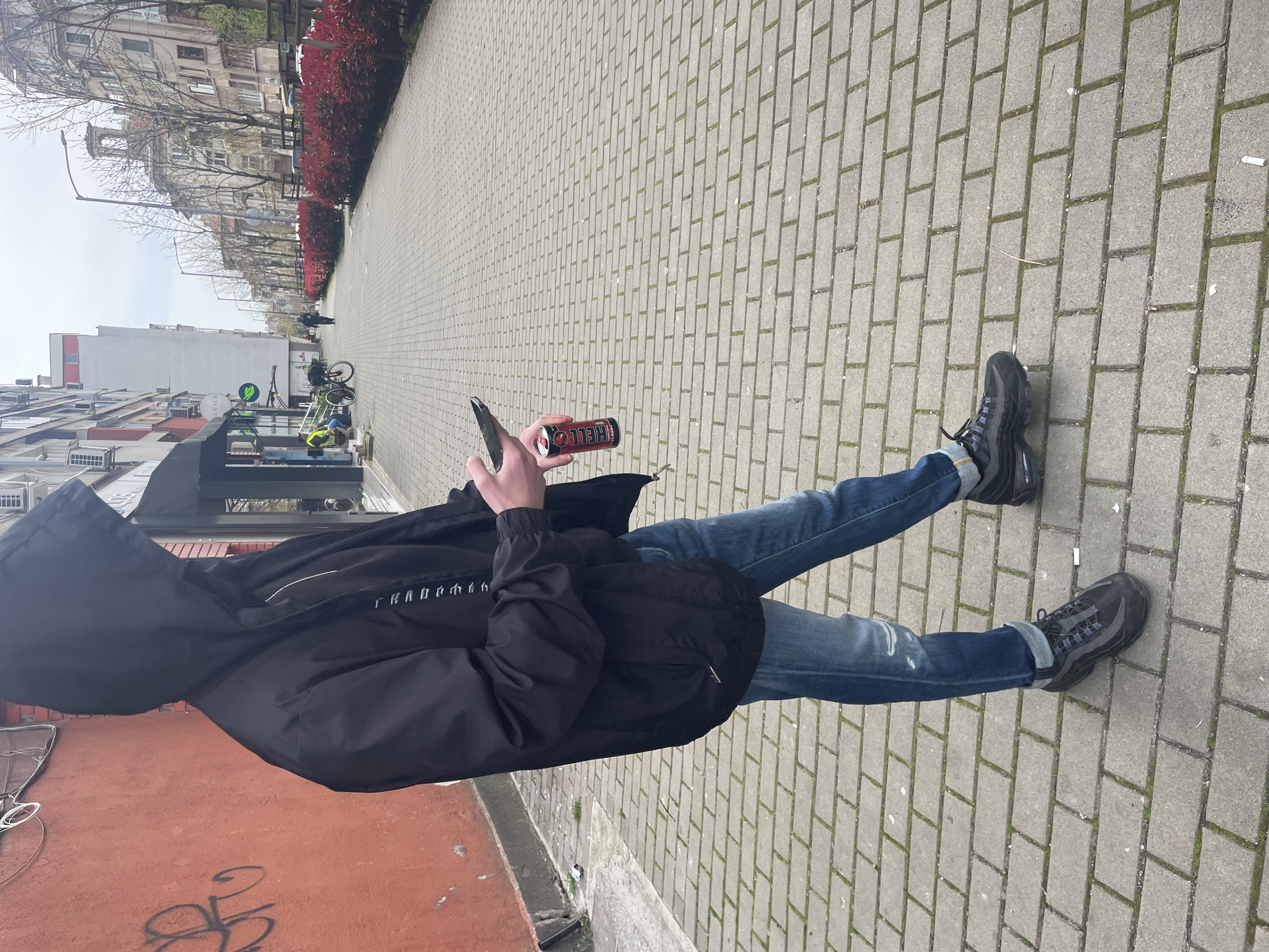 A person stands on a sidewalk in Burgas, Bulgaria. The sidewalk is paved with gray rectangular stones. Buildings line the street. A row of red-leaved bushes grows along the edge of the sidewalk. The person wears jeans, black shoes, and a black jacket with a hood. They hold a mobile phone in one hand and a red energy drink can in the other. The person appears to be looking at their phone. Graffiti is visible on a red building.