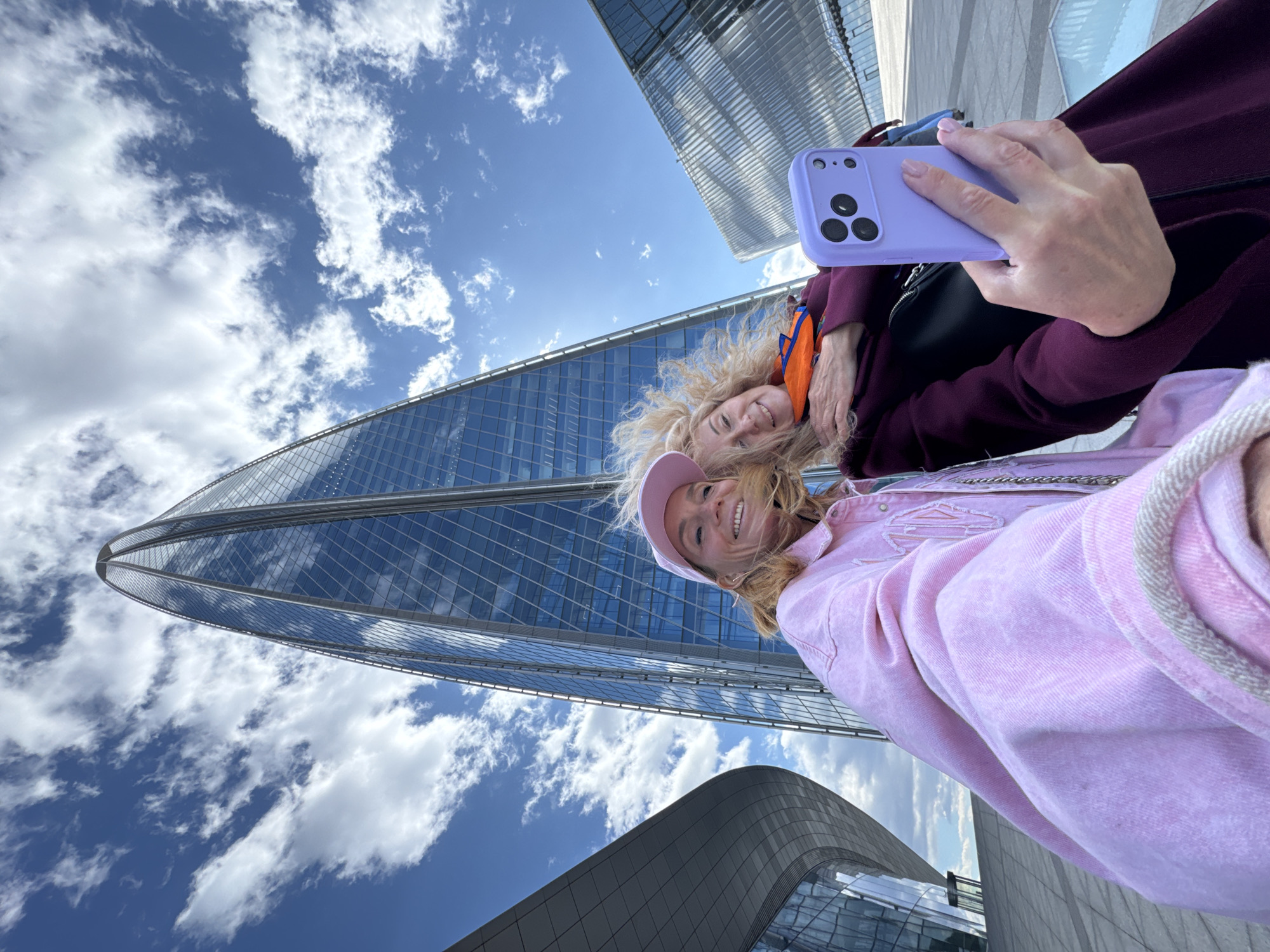 Two light-skinned women are taking a selfie with a purple smartphone in Saint Petersburg, Russia, with modern architecture and a blue sky dotted with white clouds in the background. The woman closest to the camera is wearing a light pink denim jacket and a pink cap, while the other woman is wearing a dark maroon coat with an orange strap. The photo is taken from a low angle, capturing the height and grandeur of the surrounding buildings.