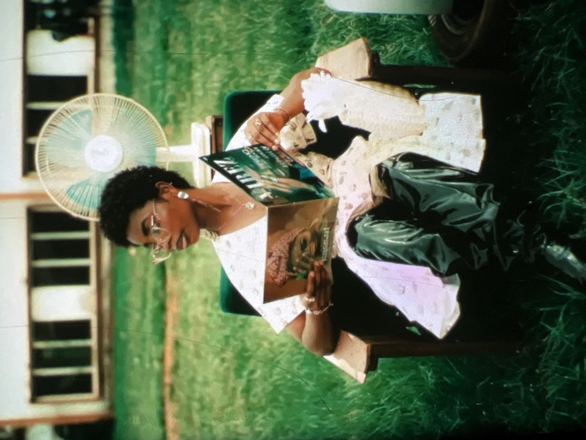 A young Black woman with short curly hair and glasses sits in a wooden chair outdoors, reading a magazine. She is wearing a white top and dark, possibly leather, pants. A white electric fan stands behind her. The background shows a building with windows and a grassy lawn, suggesting a residential or institutional setting. The scene appears to be in Lagos, Nigeria, given the context provided.