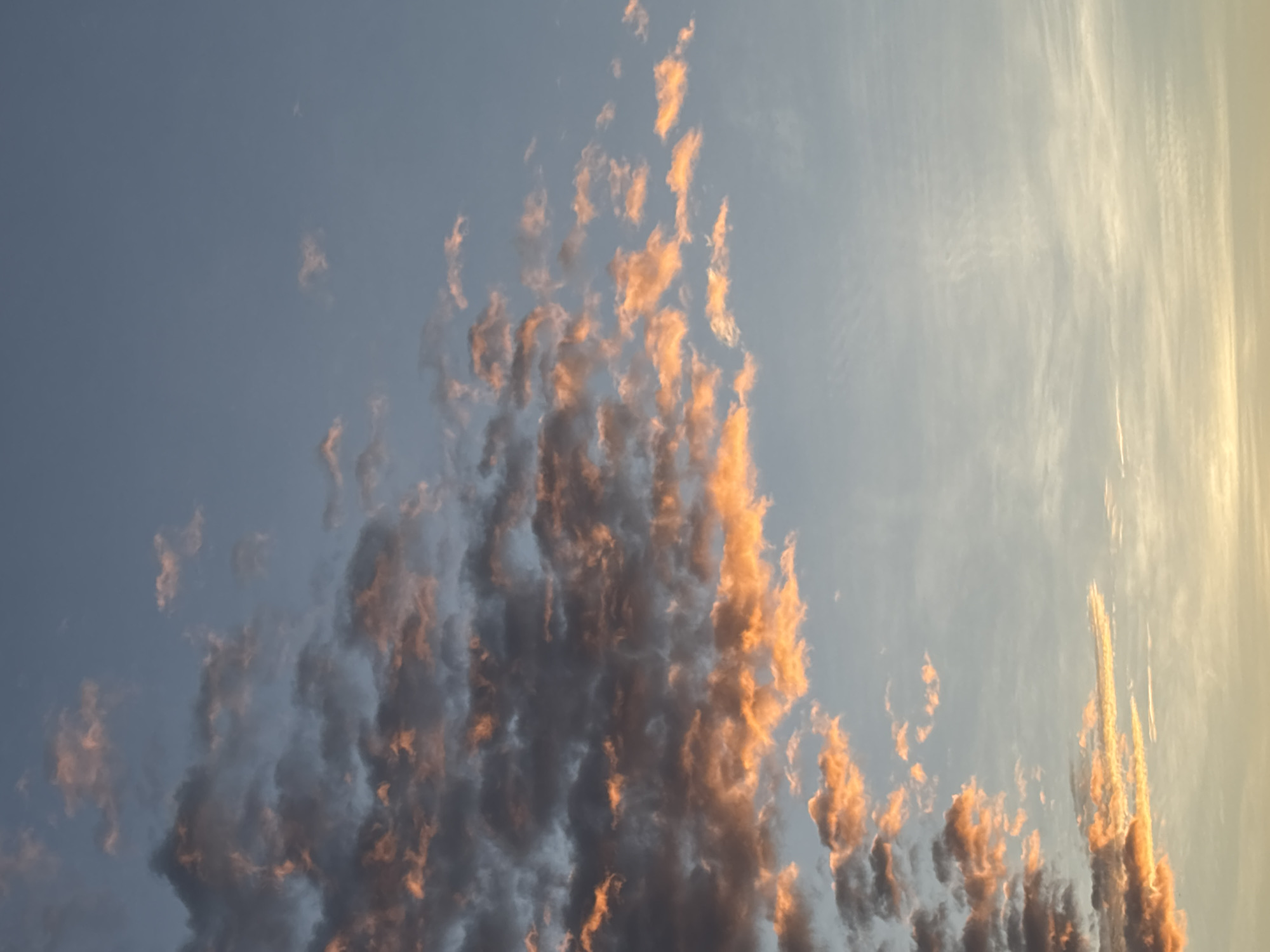 The image captures a sky filled with scattered clouds at what appears to be either sunrise or sunset. The clouds on the left side of the frame are darker, with edges illuminated by a warm, orange glow, suggesting they are catching the light of the sun. These clouds are more dense and textured. Further to the right, the clouds become thinner and more wispy, with a softer, diffused orange and white coloration. The sky itself is a gradient of pale blue at the top, transitioning to a lighter, almost yellowish hue towards the bottom horizon. There are no discernible landmarks or structures in the image, and no people or objects are visible. The scene is purely atmospheric, focusing on the interplay of light and clouds in the sky. Without any specific geographical markers within the frame, it is not possible to determine if this sky is located in Nha Trang, Vietnam, or any other specific city.