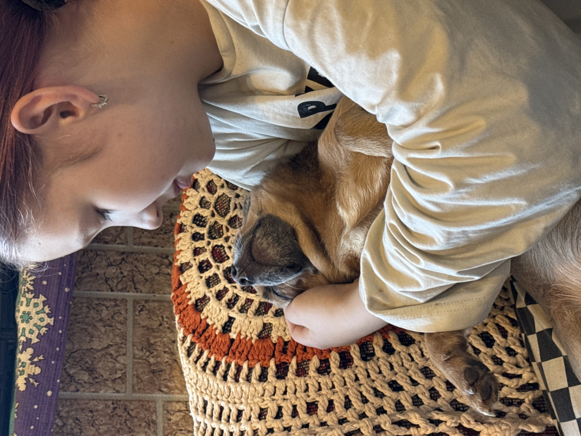 In a close-up, horizontal shot, a young girl with auburn hair pulled back holds a small, tan dog in her arms. The girl is seen from the side, looking down at the dog with a gentle expression. She is wearing a light-colored, long-sleeved shirt with a black graphic on it. The dog is nestled in her arms, its eyes closed and head tilted back, appearing to be asleep or resting contentedly. The dog has a tan coat and grey muzzle.
The girl and dog are resting on a textured, crocheted surface with a pattern of cream, orange, and darker colors. Behind them, a textured, brown wall is visible, possibly a brick or stone surface, with a decorative purple fabric draped above it. The background is slightly out of focus, drawing attention to the subjects. The scene suggests a moment of quiet affection and comfort. No information is provided to indicate a specific location in Moscow, Russia.