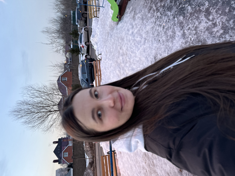 The scene is an outdoor environment in Akulovo (Gp Kubinka), Russia, characterized by patchy snow and ice cover on the ground. A female subject occupies the foreground, facing the camera. She presents with long, dark brown hair and is dressed in a dark jacket over a white hooded sweatshirt. Her facial expression is a slight smile, with her eyes directed slightly left of the camera's axis. To the subject's left, a portion of a wooden slatted bench with blue metal supports is visible. To her right, part of a green plastic playground slide is observed. In the midground, a paved area serves as a parking space for several vehicles, including a dark SUV and a light-colored sedan. The background features a low earth-toned retaining wall, beyond which are multiple detached residential structures with pitched roofs and numerous bare deciduous trees. The sky is pale blue, suggesting ambient light conditions consistent with dawn or dusk.