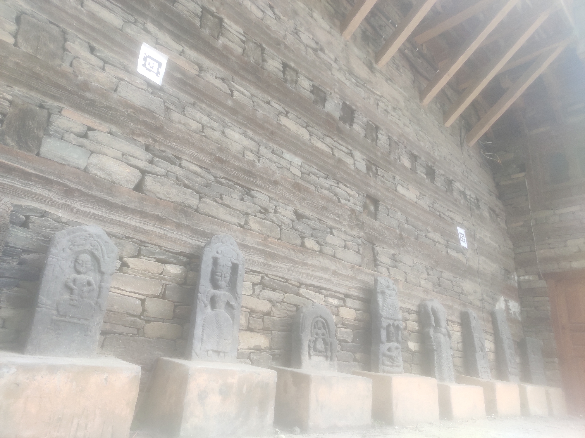 The image shows a stone wall with several stone carvings of figures on display, set on square pedestals. The wall is constructed of roughly hewn stones and interspersed with horizontal wooden beams. Above the wall, wooden rafters form a sloping roof. In the upper left of the image, a square white marker with a QR code is affixed to the wall. The entire scene is slightly overexposed, giving it a hazy appearance. The image is likely taken in Naggar, India, possibly at a historical or cultural site showcasing ancient sculptures.