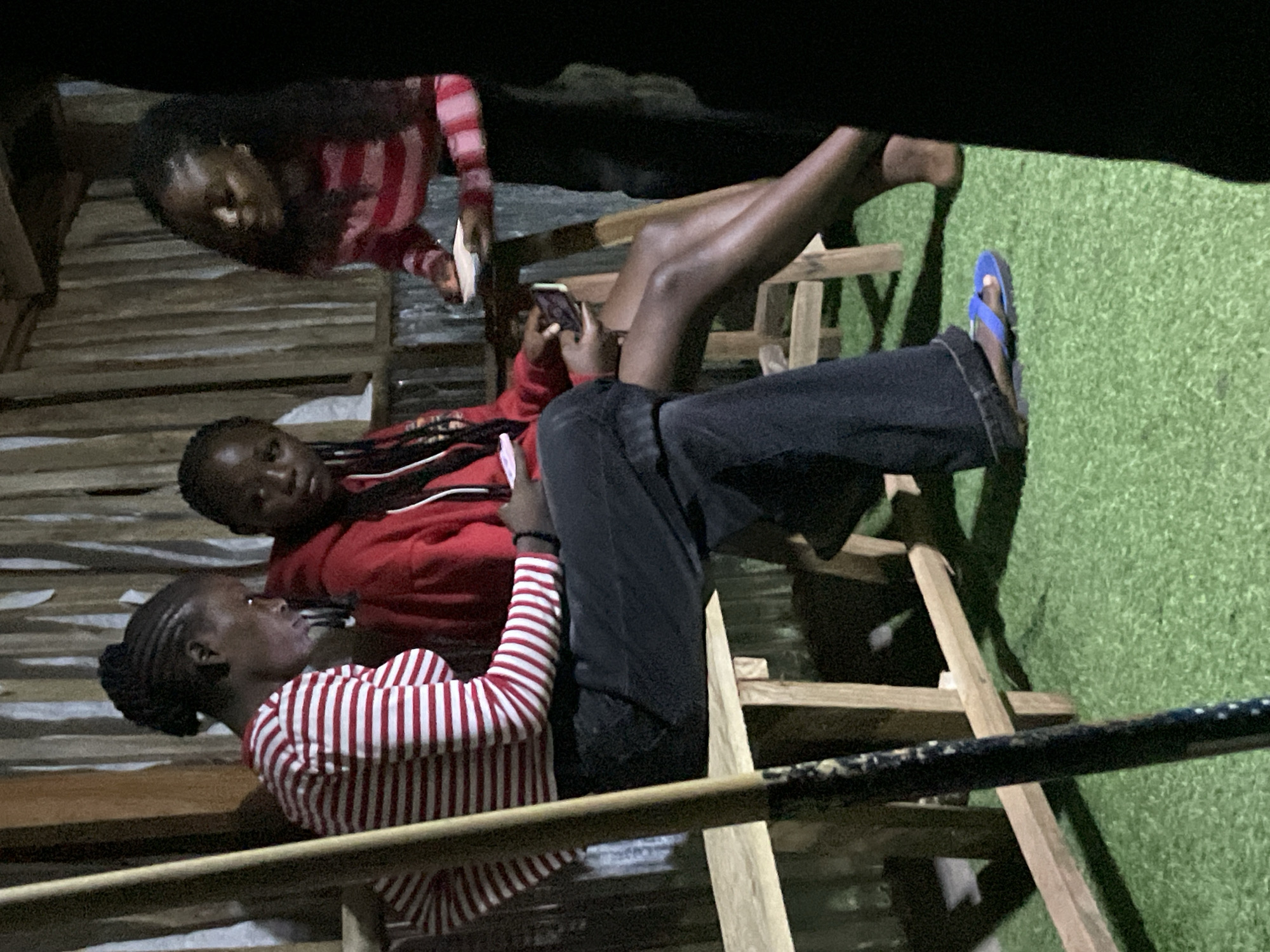 In Durumi, Nigeria, a group of three young Black individuals, two females and one other whose gender is not fully discernible due to cropping, are gathered in a dimly lit, outdoor setting. The environment appears to be rustic, with vertical wooden planks forming a backdrop and a corrugated metal surface visible behind them. Artificial turf covers the ground.
The two prominent females are seated on a wooden bench. The female on the left wears a red and white horizontally striped long-sleeved shirt and dark pants, with her hair styled in braids pulled back. She is looking down, engaged with a mobile phone held in her hands. The female in the center is wearing a red hooded sweatshirt and dark pants, also looking down, potentially at a device. Her hair is in braids that trail down her shoulders.
To the right, a third individual, partially visible and cropped, is standing and holding a small white object, possibly a plate or cup, and looking towards the seated individuals. This individual is wearing a red and white horizontally striped long-sleeved shirt, similar in pattern to the person on the left.
A wooden pole, possibly part of a larger structure or support, is positioned prominently in the left foreground, out of focus. The overall lighting suggests the scene takes place after dusk.