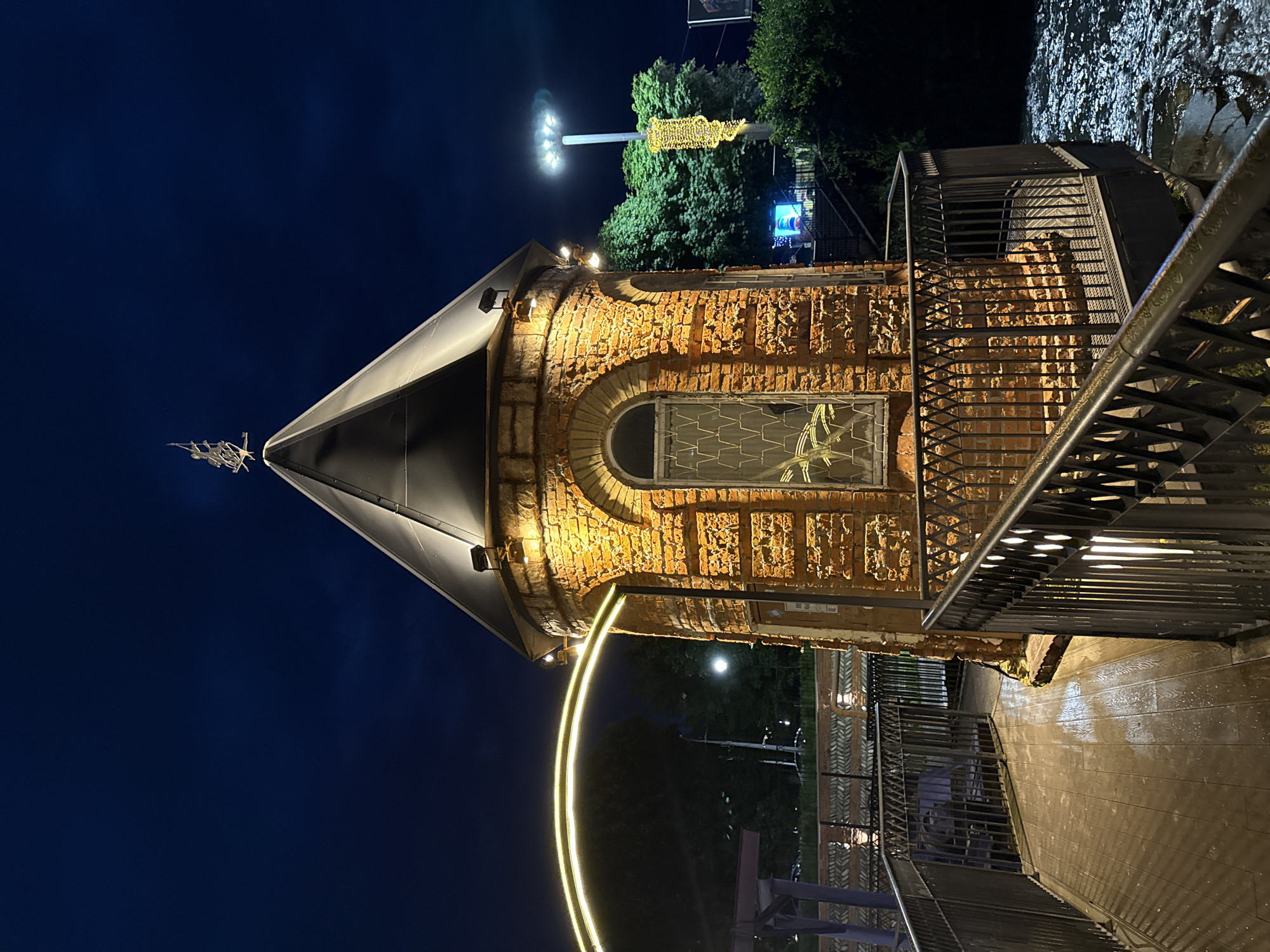 The image depicts a nighttime scene in Vladikavkaz, Russia. The primary subject is a small, brick tower with a dark conical roof, illuminated by warm artificial lights. The tower is situated near a body of water, possibly a river, with a wet, wooden walkway in the foreground. A metal staircase with railings leads up to the tower.
In the background, trees are visible, along with streetlights and a decorative light installation resembling a string of lights on a pole, creating a festive atmosphere. The sky is dark, indicating it is nighttime, and the wet surfaces suggest recent rainfall or high humidity. There are no people visible in the image. The overall mood is serene and somewhat magical, enhanced by the nighttime lighting. The scene appears to be an outdoor public space, possibly a park or waterfront area. No visible text is present in the image.