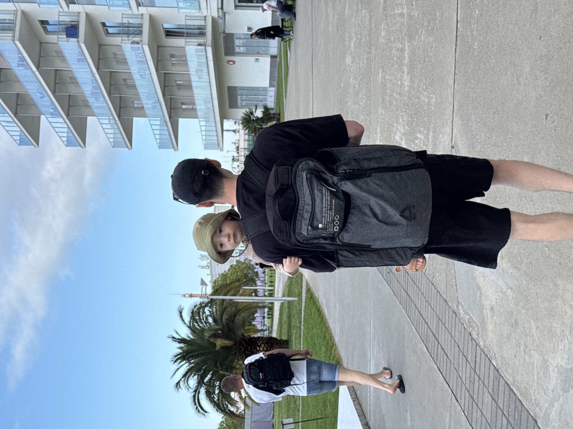 A man, seen from the back, carries a small child on his back. The man is wearing a black baseball cap, a black t-shirt, and black shorts. A grey backpack is on his back, partially obscuring the child, who is wearing a beige hat. They are on a paved area next to a patch of green grass with palm trees. In the background, there is a large modern building with stacked balconies. Another adult is walking in the grass, and other adults are visible in the distance. The image was taken outside on a bright day. The environment suggests a city setting, likely a resort or tourist area. Given the visual cues, the location is likely Sochi, Russia.
