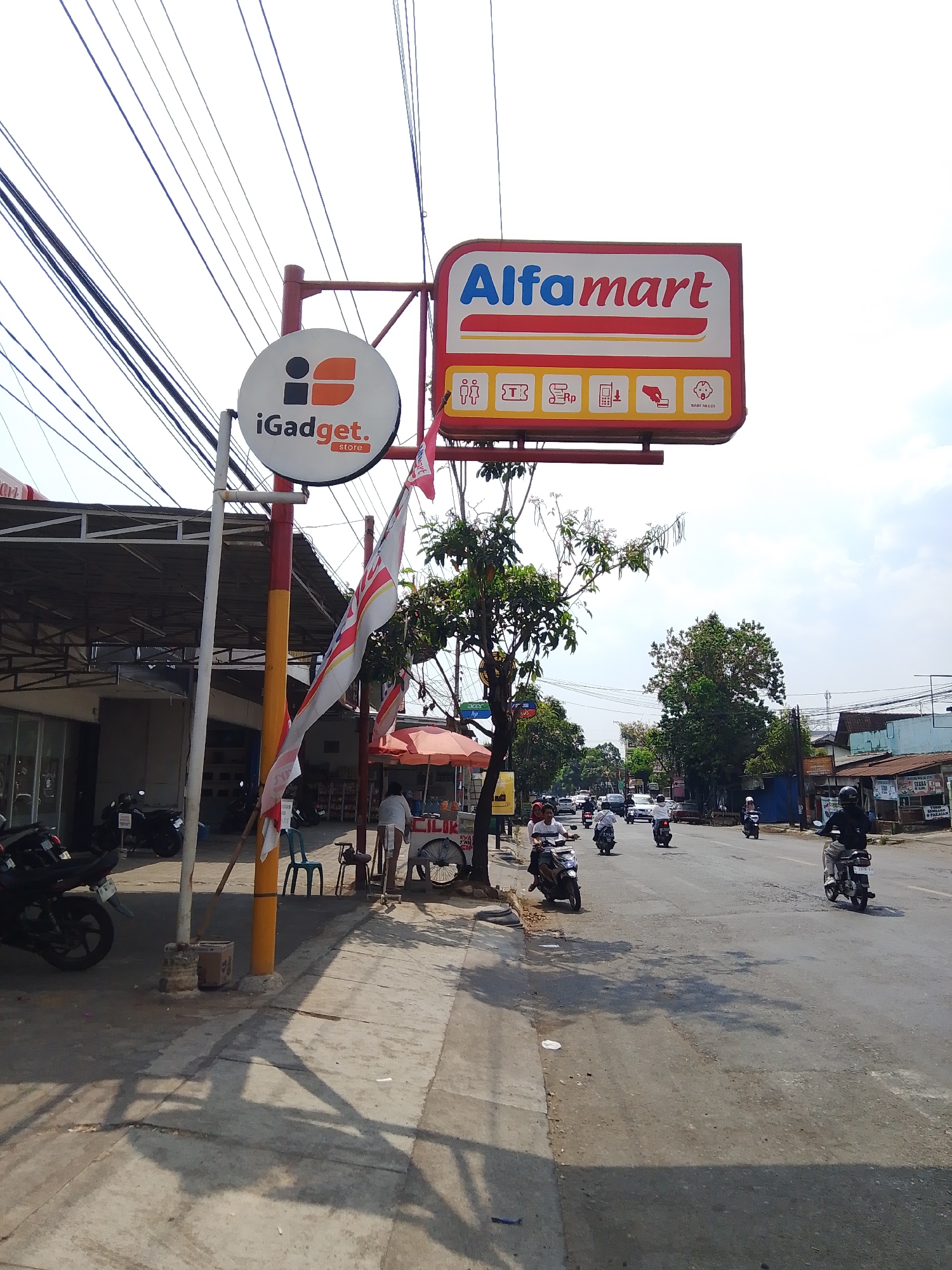 This is an eye-level, outdoor, medium-shot photograph taken on a street in Potroyudan, Indonesia. The image captures a street scene with a focus on commercial signage and passing traffic.

Dominating the upper portion of the image is a red and white sign for "Alfamart," a convenience store chain, featuring its logo and several icons indicating services such as "baby needs." Below the Alfamart sign, and slightly to the left, is a circular white sign for "iGadget. store" with a stylized orange and black logo.

The street itself is paved and appears to be a main thoroughfare. Multiple motorcycles are visible, some parked along the side of the road and others in motion. One motorcycle is in the foreground on the left, and several more are seen further down the street, some with riders. Cars are also present in the background, indicating traffic flow.

On the sidewalk to the right of the signs, there are a few people, one standing near a food cart with an umbrella, and another person sitting on a chair. A woman in a white shirt and shorts is visible standing near the food cart. A flag, possibly the Indonesian flag given its red and white colors and design, is attached to a pole near the iGadget sign and extends outwards.

The environment includes some trees and buildings lining the street. Overhead, a complex network of power lines stretches across the sky, which is partly cloudy. The lighting suggests it is daytime, with shadows cast on the ground, indicating the sun is present. The overall scene depicts a busy commercial area in an Indonesian city.