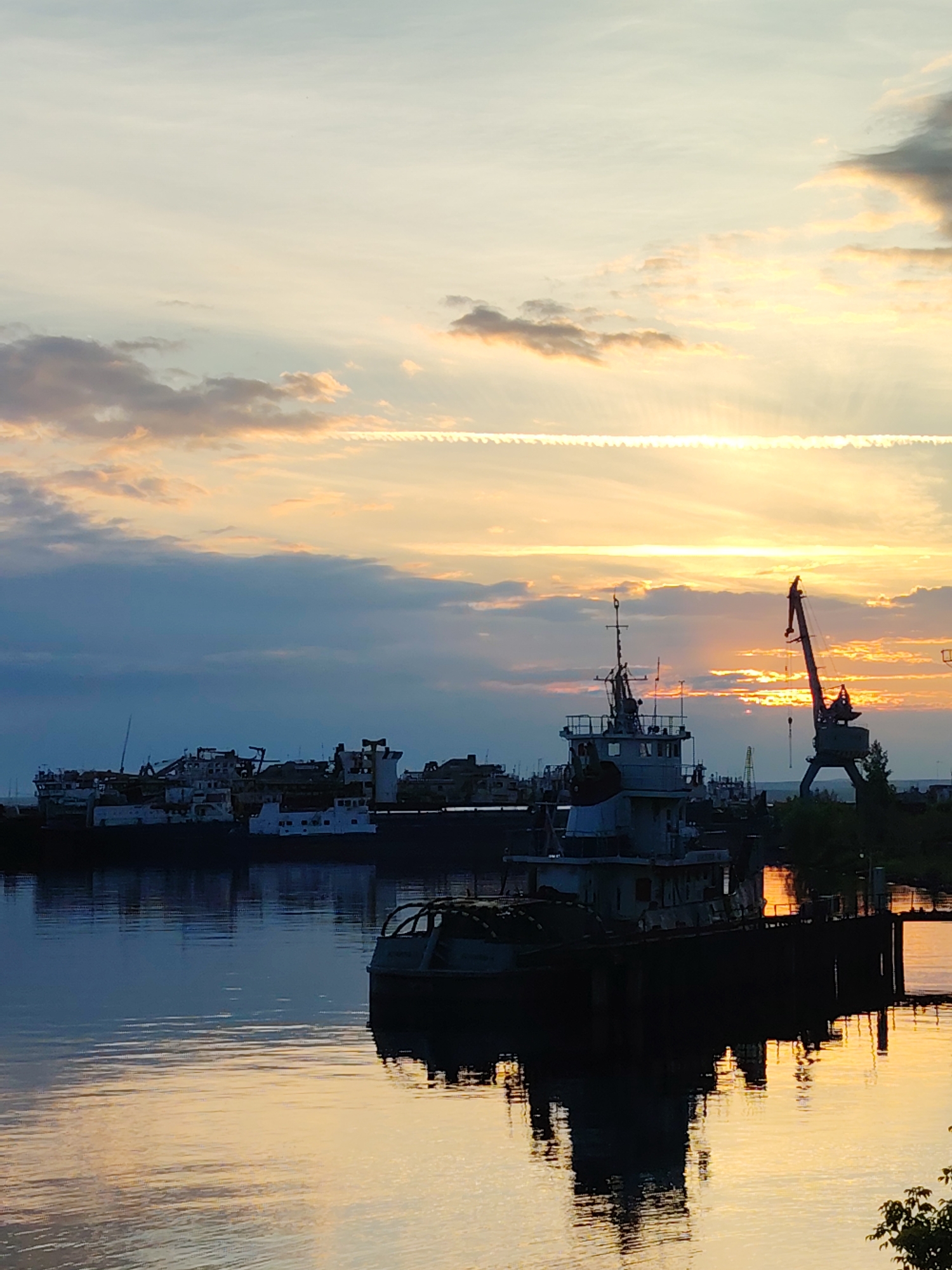 The image depicts a view of the Chaikovsky, Russia port area at sunset. In the foreground, the calm water of the Kama River reflects the warm hues of the sky and the silhouettes of several boats and industrial equipment. A tugboat is prominently featured in the middle ground, partially submerged and casting a dark reflection on the water. In the background, a line of various ships and barges are moored along the docks, their forms softened by the fading light. A large construction crane stands tall on the right side of the image, its silhouette sharp against the vibrant orange and yellow sky. Clouds are scattered across the sky, some catching the sun's rays and appearing as streaks of light. The overall scene conveys a sense of industrial activity at the end of the day, with the natural beauty of the sunset adding a dramatic backdrop.