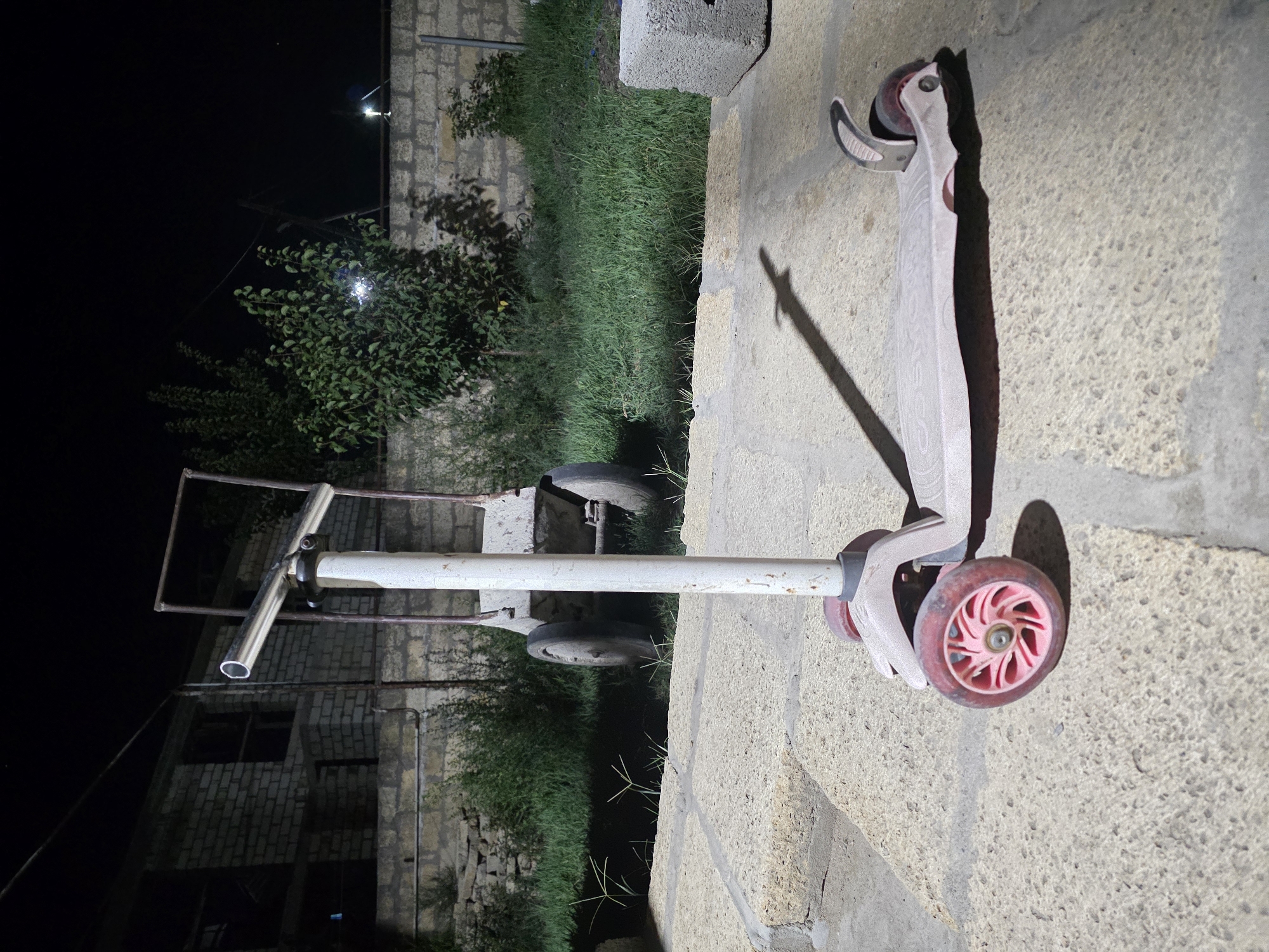 A pink, three-wheeled scooter is positioned in the foreground on a paved surface at night. The scooter is facing to the right, with its handlebars extending upwards and to the left. Behind the scooter, a cart with two wheels is visible, as well as a brick building and some vegetation. The scene is illuminated by artificial light, casting shadows. The overall environment appears to be outdoors, possibly a yard or an alleyway.